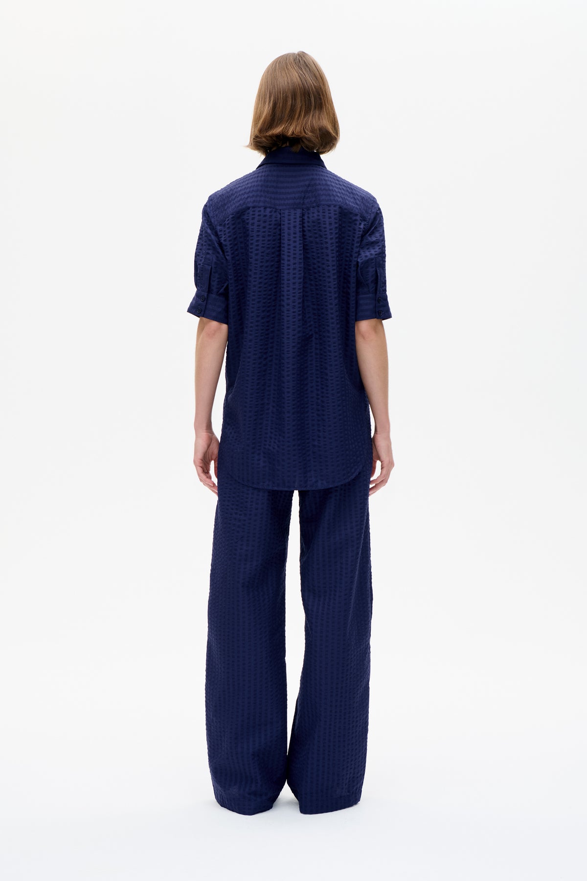 Back view of a model wearing a navy short-sleeved collared shirt and matching wide-leg trousers with a subtle raised pattern.