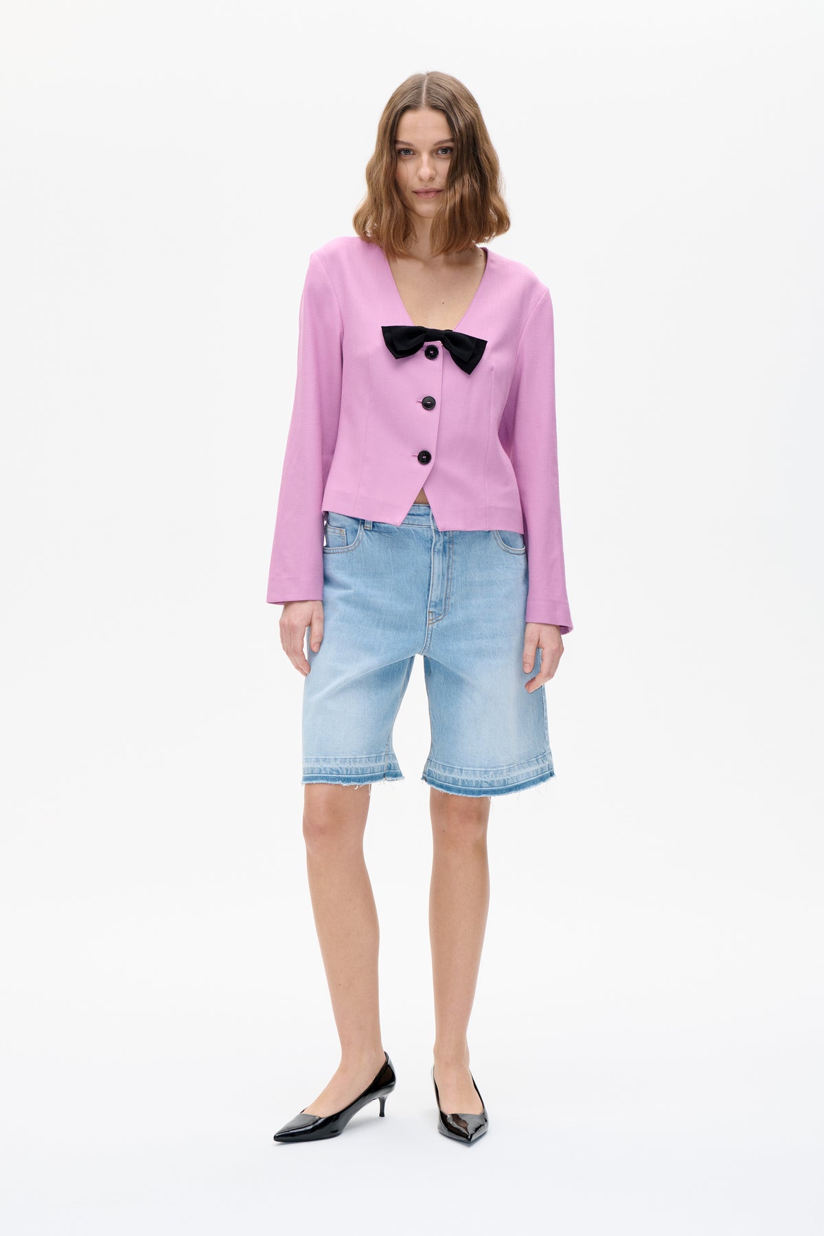Vorderansicht eines Models, das eine rosa Jacke mit V‑Ausschnitt, einer schwarzen Schleife und schwarzen Knöpfen trägt, kombiniert mit hellen denim-Shorts.