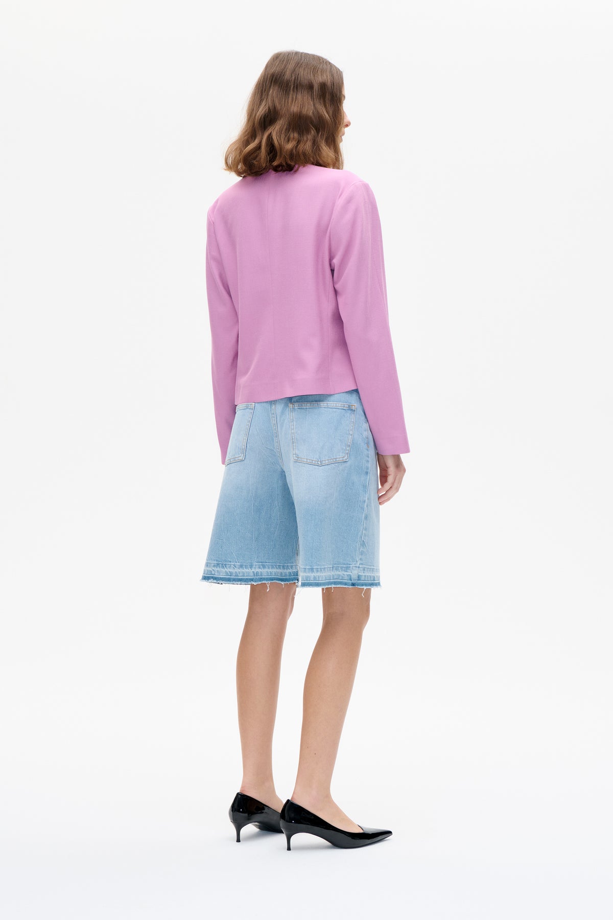 Rückenansicht eines Models mit schlichtem fliederfarbenem Langarmtop, hell gewaschenen Denim-Shorts und schwarzen spitzen High Heels.