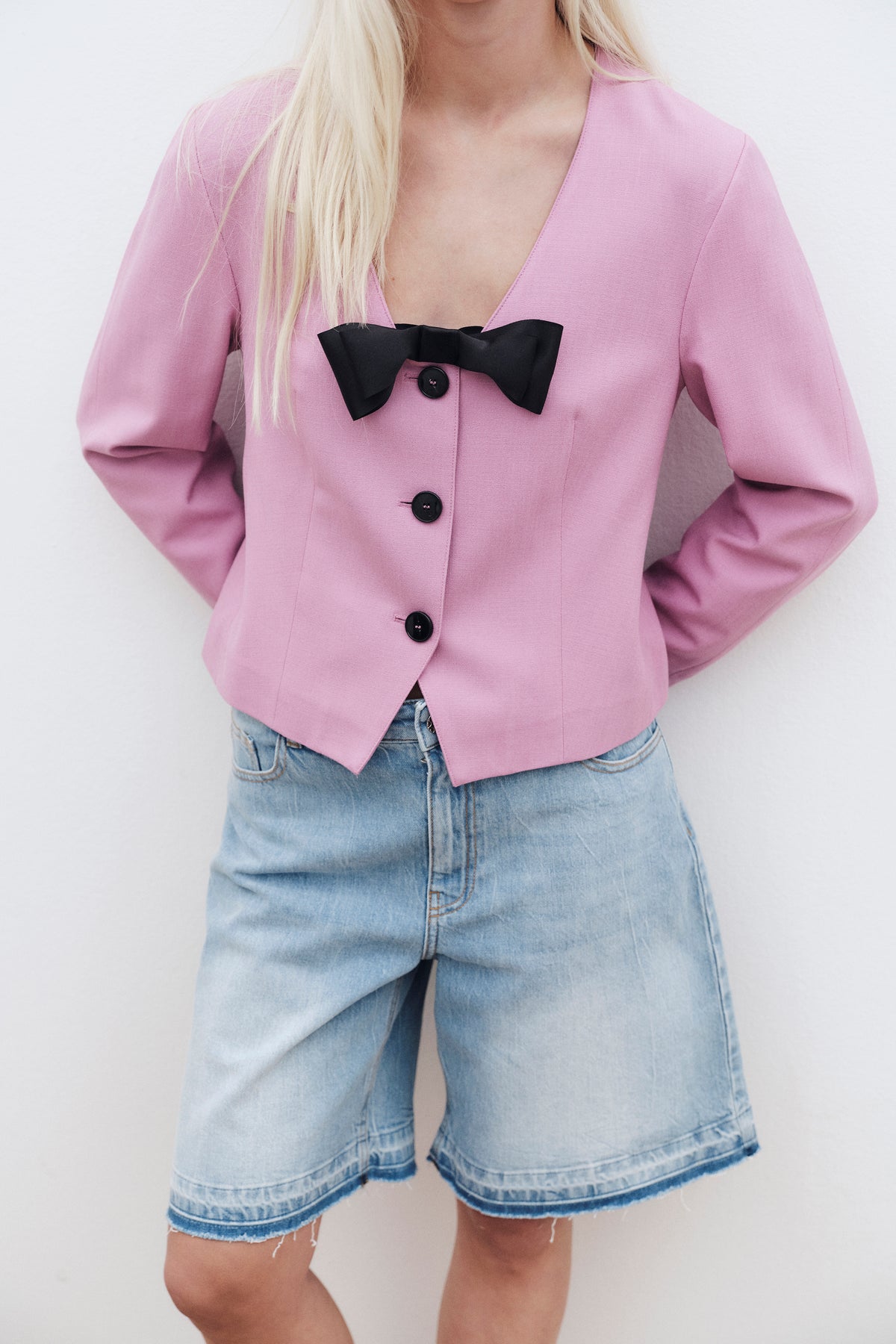 Nahaufnahme aus der Kampagne: Model trägt pinken, taillierten Blazer mit schwarzer Schleife und Knöpfen, kombiniert mit hellen Denim-Shorts.