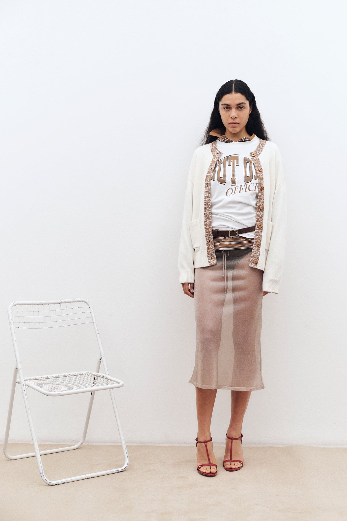 Campaign portrait with a model beside a white folding chair, wearing a cream cardigan with brown trim, graphic T-shirt, striped neck scarf and taupe skirt.