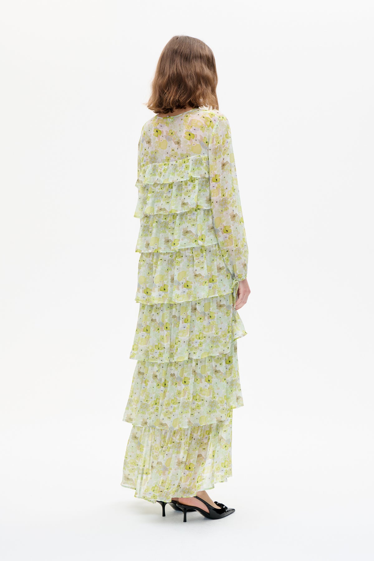 Back view of a model wearing a tiered, ruffled dress with a pale yellow and green floral print and long sleeves.
