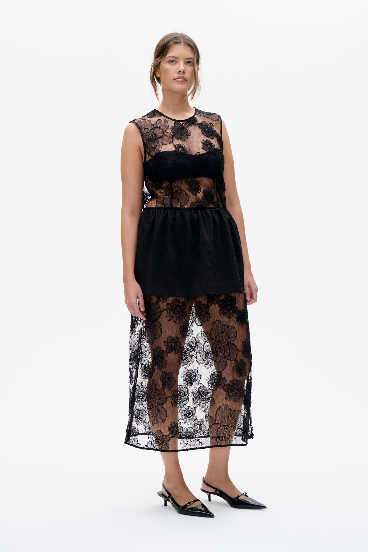 Model wearing a black floral lace midi dress with a sheer sleeveless bodice, opaque bust panel and short black skirt layer, front view