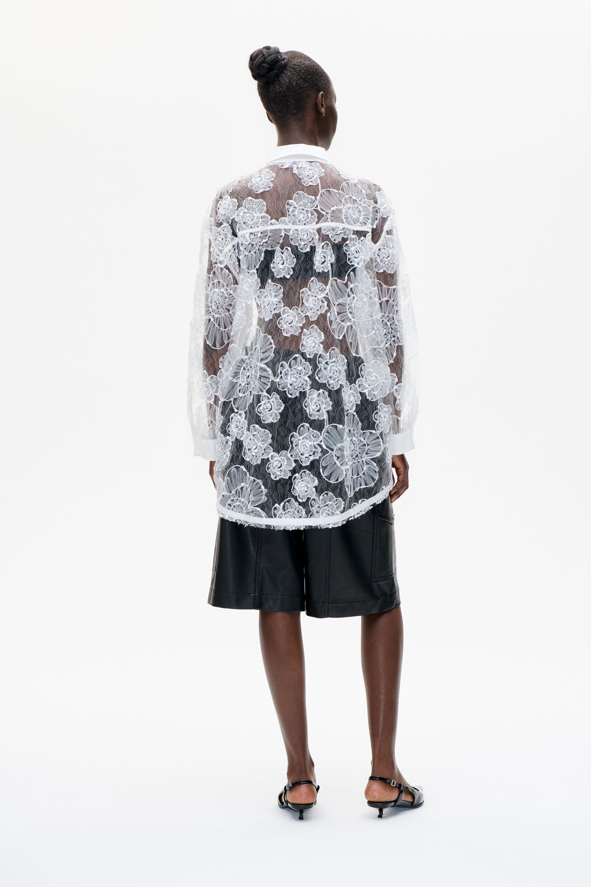 Rückansicht einer transparenten weißen Spitzenbluse mit Blumenmuster, blickdichtem weißem Kragen und Manschetten, über schwarzen Shorts getragen.