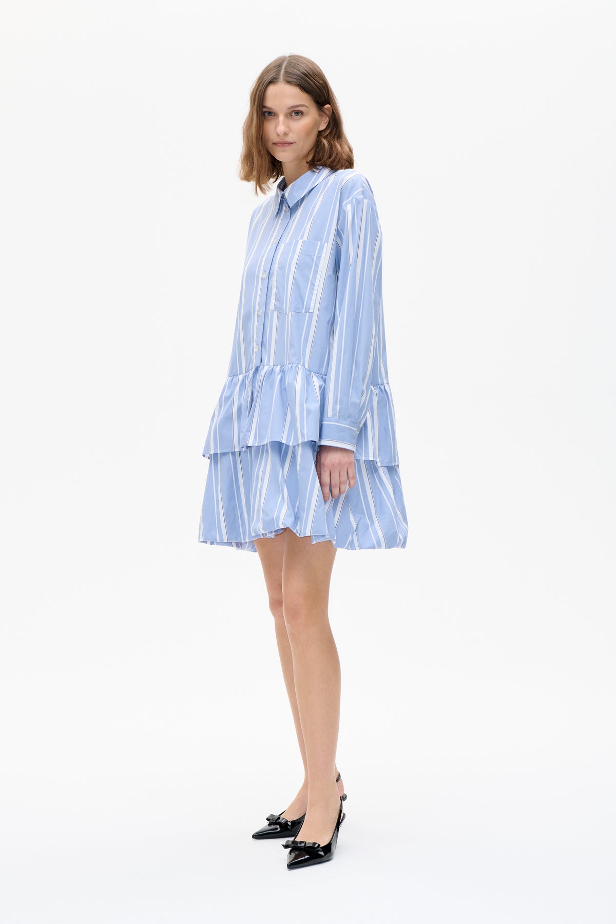 Front view of a light blue shirt dress with varied white vertical stripes, a button front, chest pocket, long sleeves and gathered flounce hem
