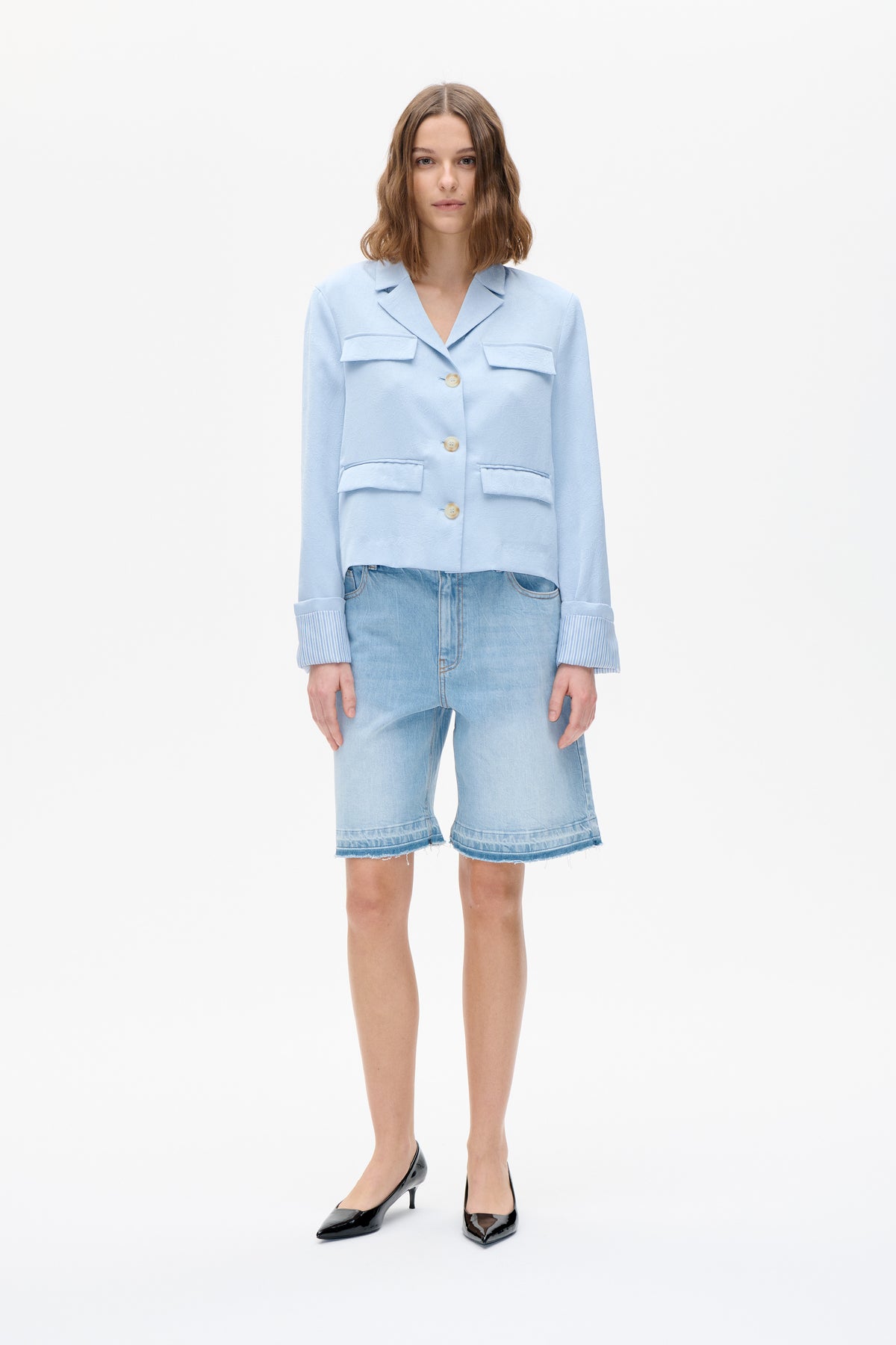 Frontalansicht eines Models, das eine hellblaue, strukturierte Jacke mit durchgehender Knopfleiste, vier Klappentaschen und gestreiften Bündchen trägt, kombiniert mit denim-Shorts.