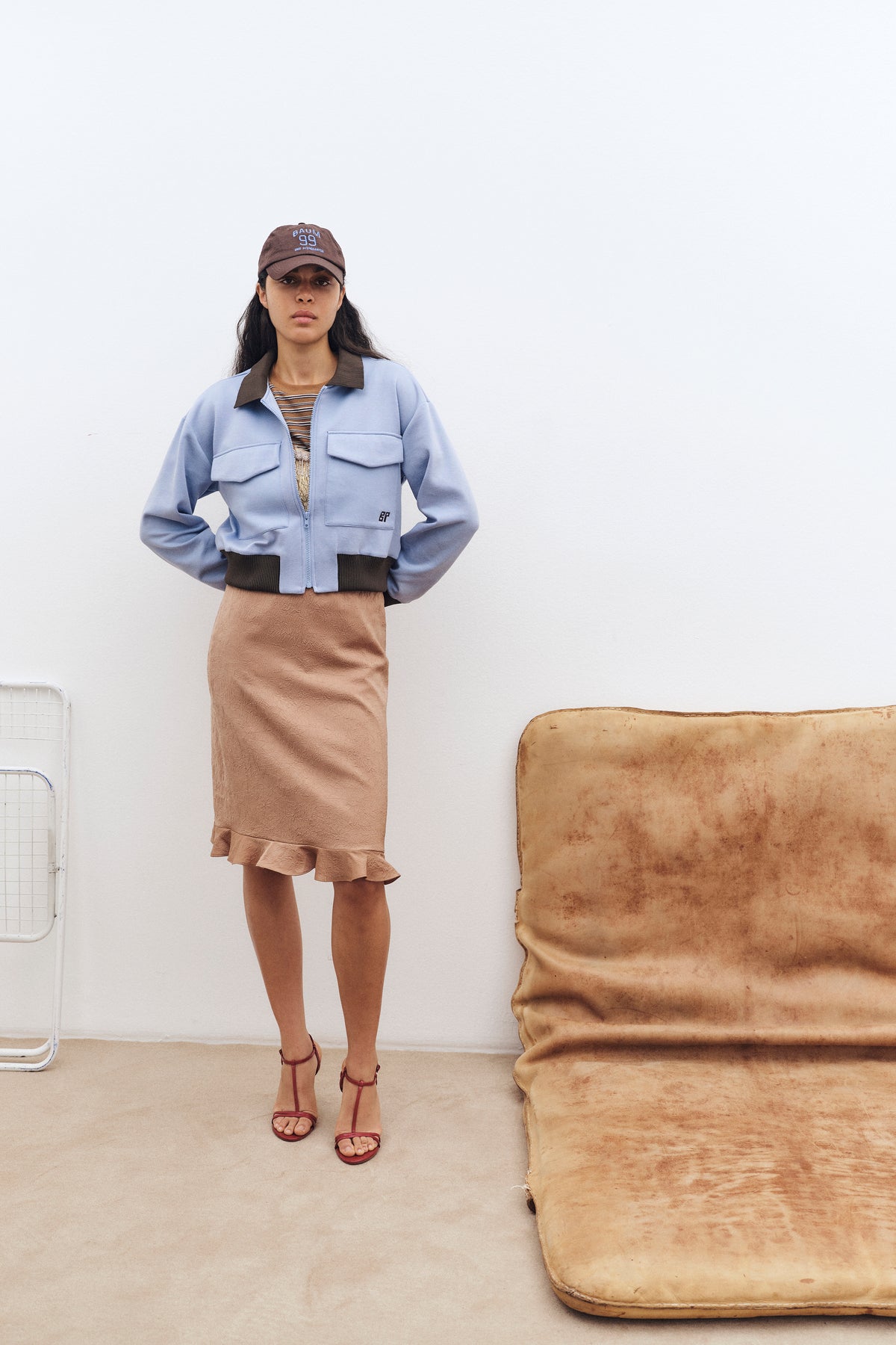 Model standing against a pale wall wears a pale blue zip-front jacket with brown trim, a striped top, textured tan midi skirt and brown cap.