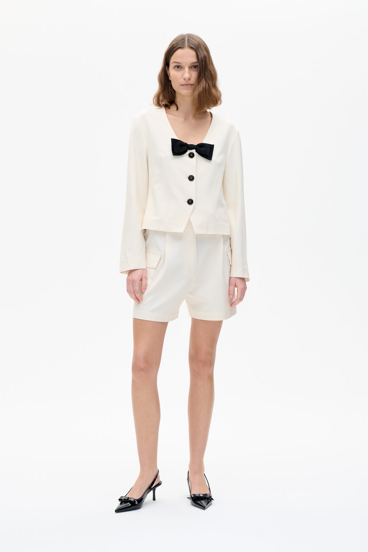 Model wearing a cream collarless jacket with black bow and buttons, matching tailored shorts, and black pointed heels