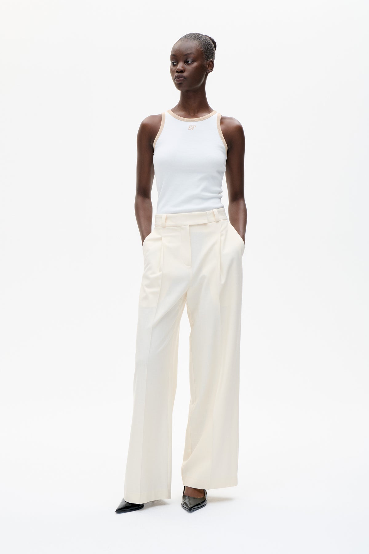 Model wearing a white sleeveless top with beige trim and a small chest monogram, paired with cream wide-leg trousers.