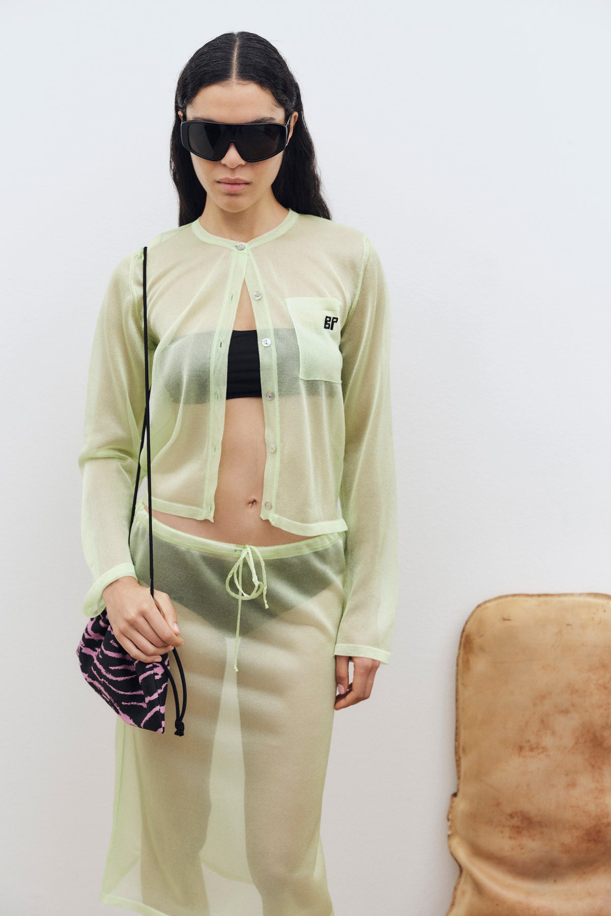 Model wearing a sheer pale-green button-front cardigan over a black crop top, with matching drawstring trousers and oversized black sunglasses.