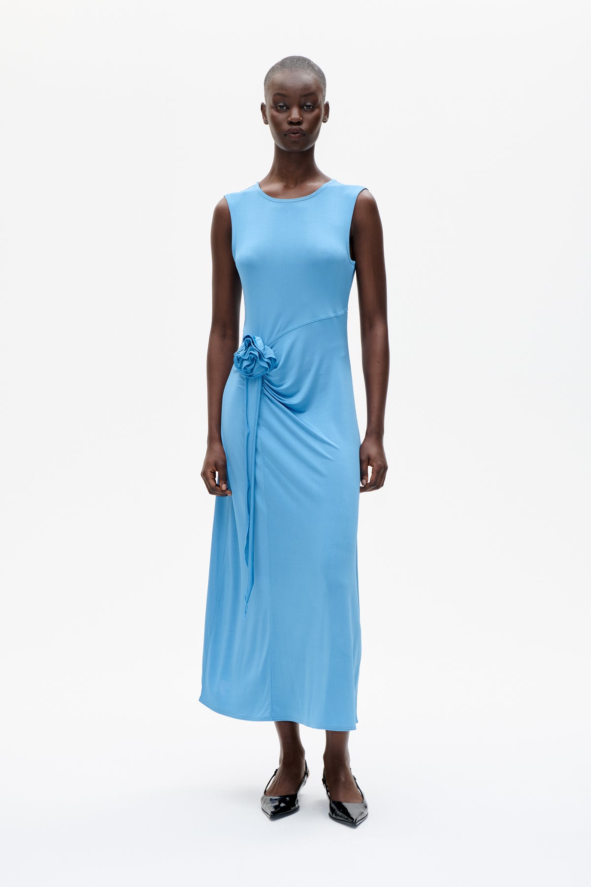 Front view of a model wearing a sky-blue sleeveless midi dress with an asymmetrical gathered waist and fabric rosette detail.