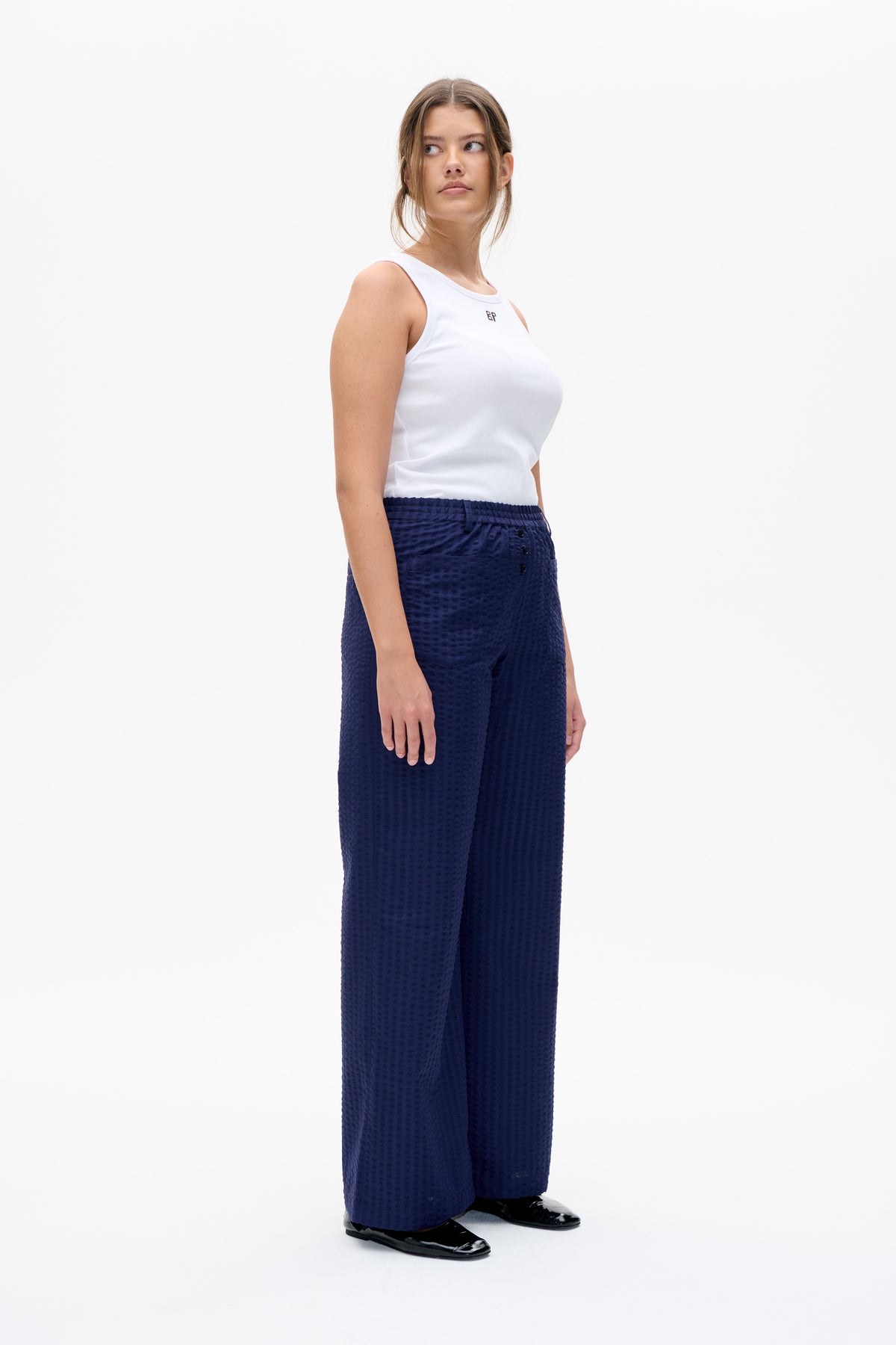 Model wearing a white sleeveless top with a small chest emblem and textured navy wide-leg trousers, shown from the front.