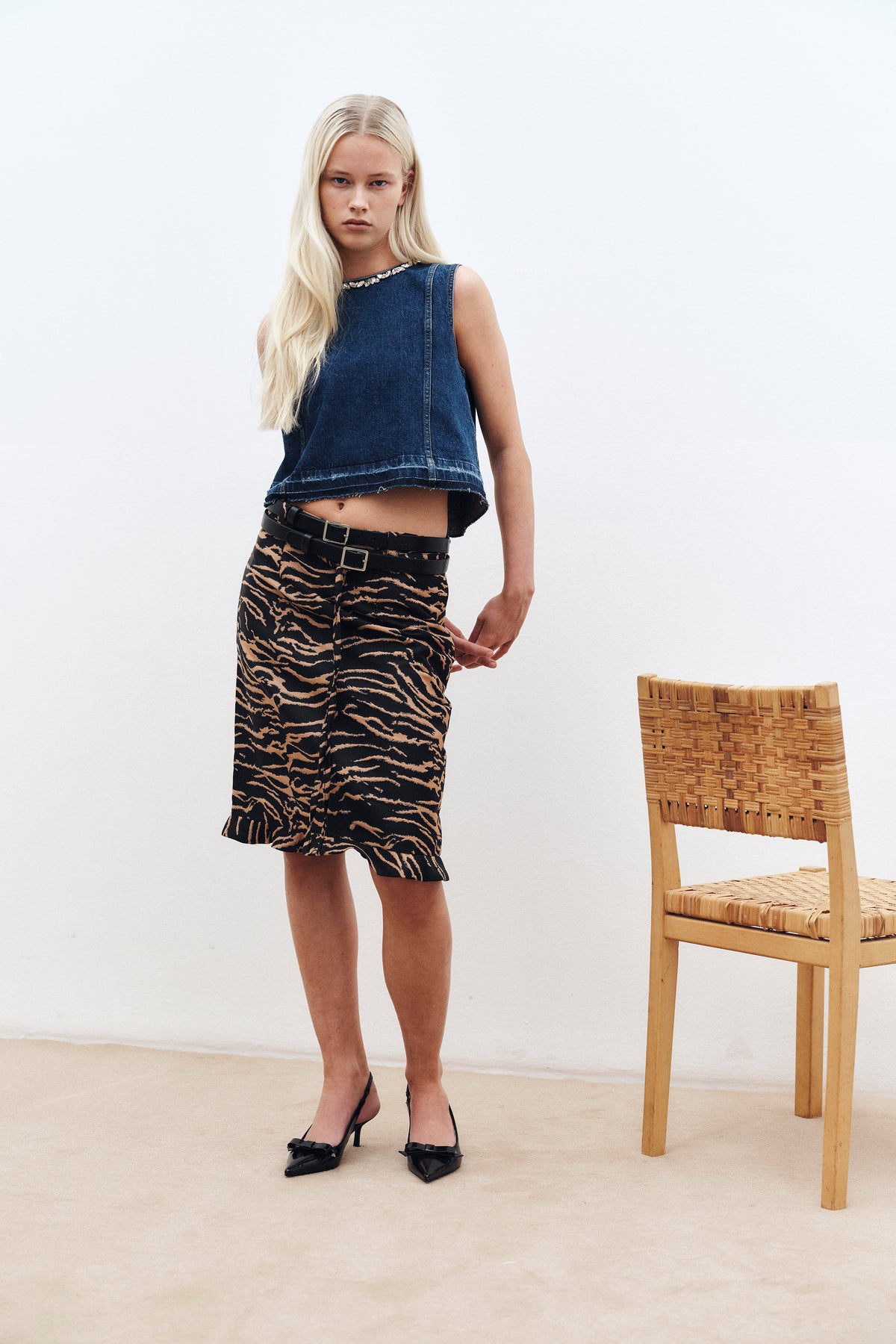 Campaign portrait of a model in a dark blue sleeveless denim top with a jeweled neckline and black-and-tan patterned skirt beside a woven chair.