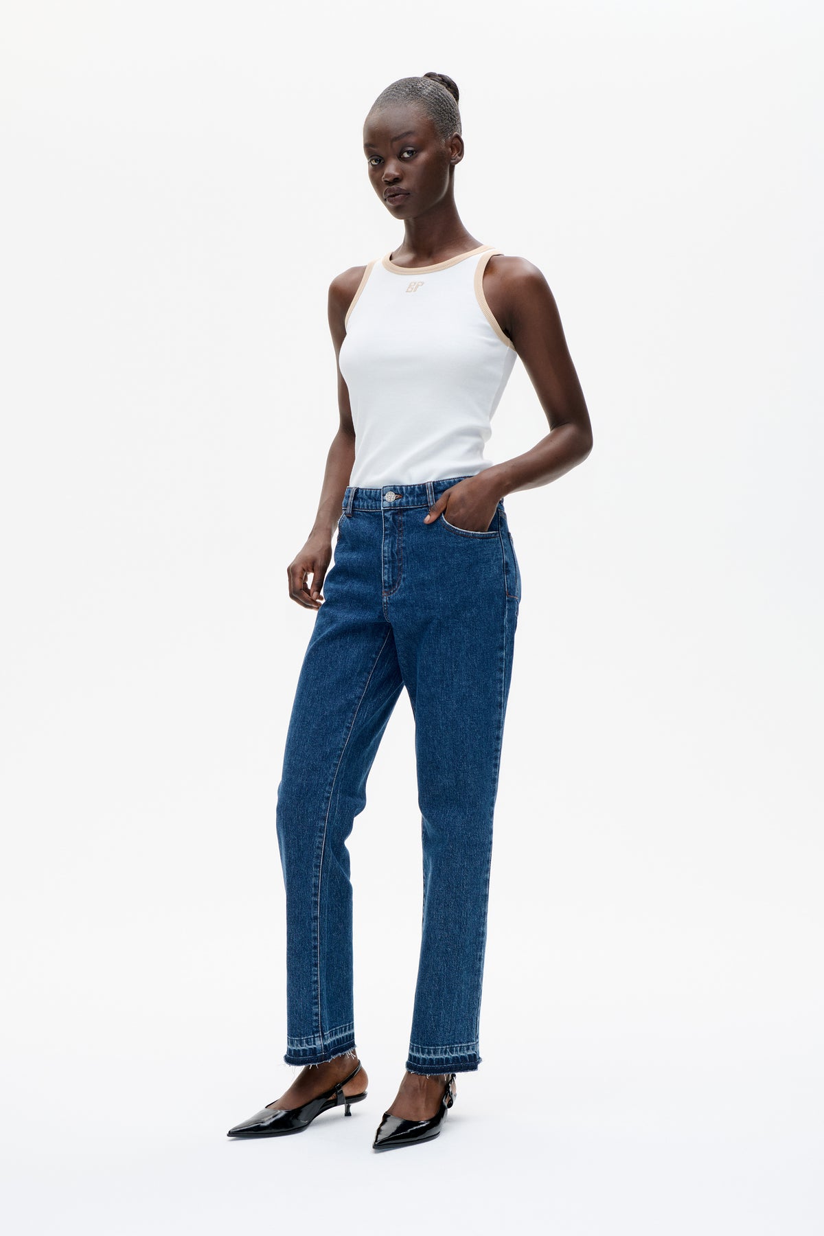 Model wearing a white sleeveless top with beige trim and blue straight-leg jeans with frayed hems, styled with black heels.