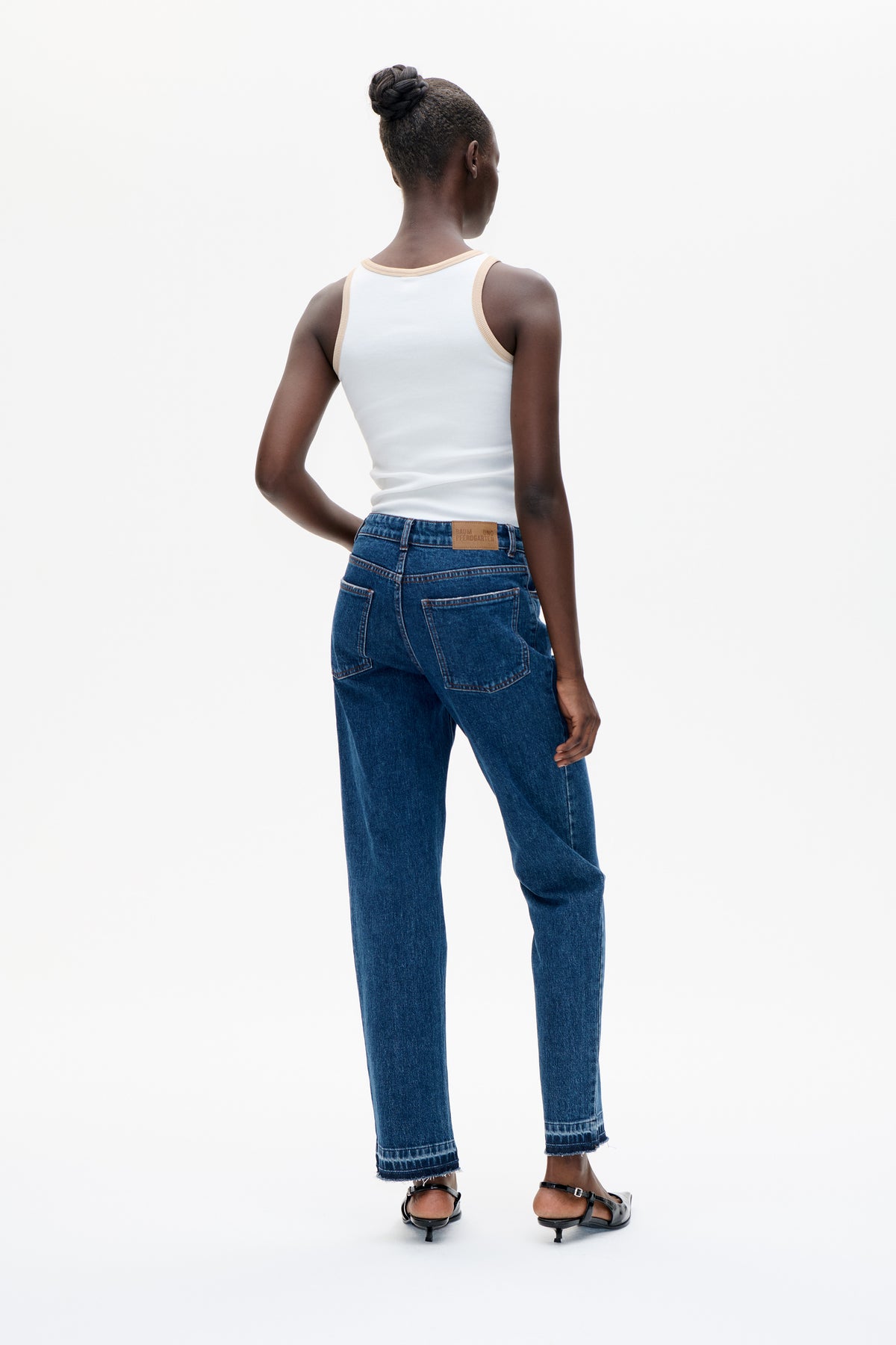Rückenansicht eines Models, das ein weißes Tanktop mit beigefarbener Einfassung und dunkelblaue, verkürzte Jeans mit Gesäßtaschen und rohen Säumen trägt.