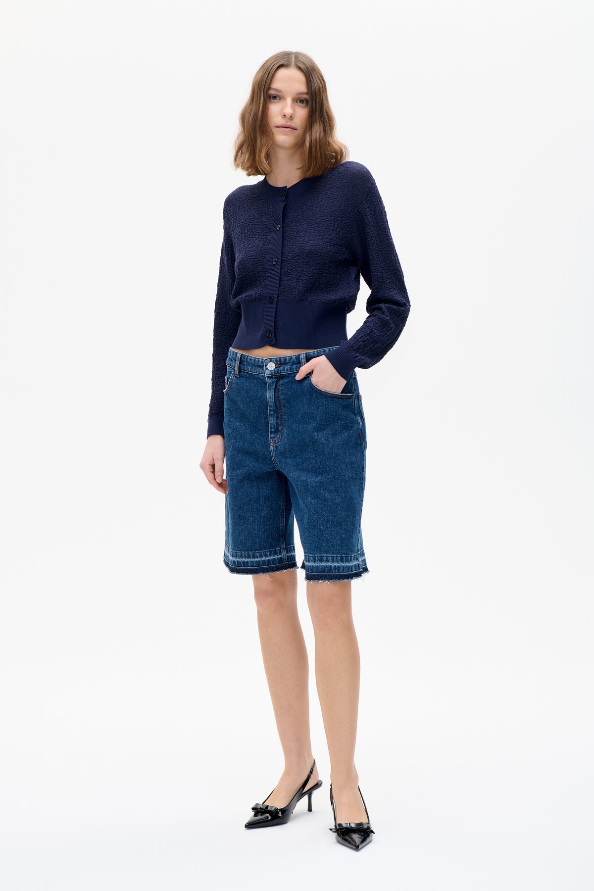 Model wearing a navy textured button-front cardigan with blue denim knee-length shorts and black bow-detail slingback heels.