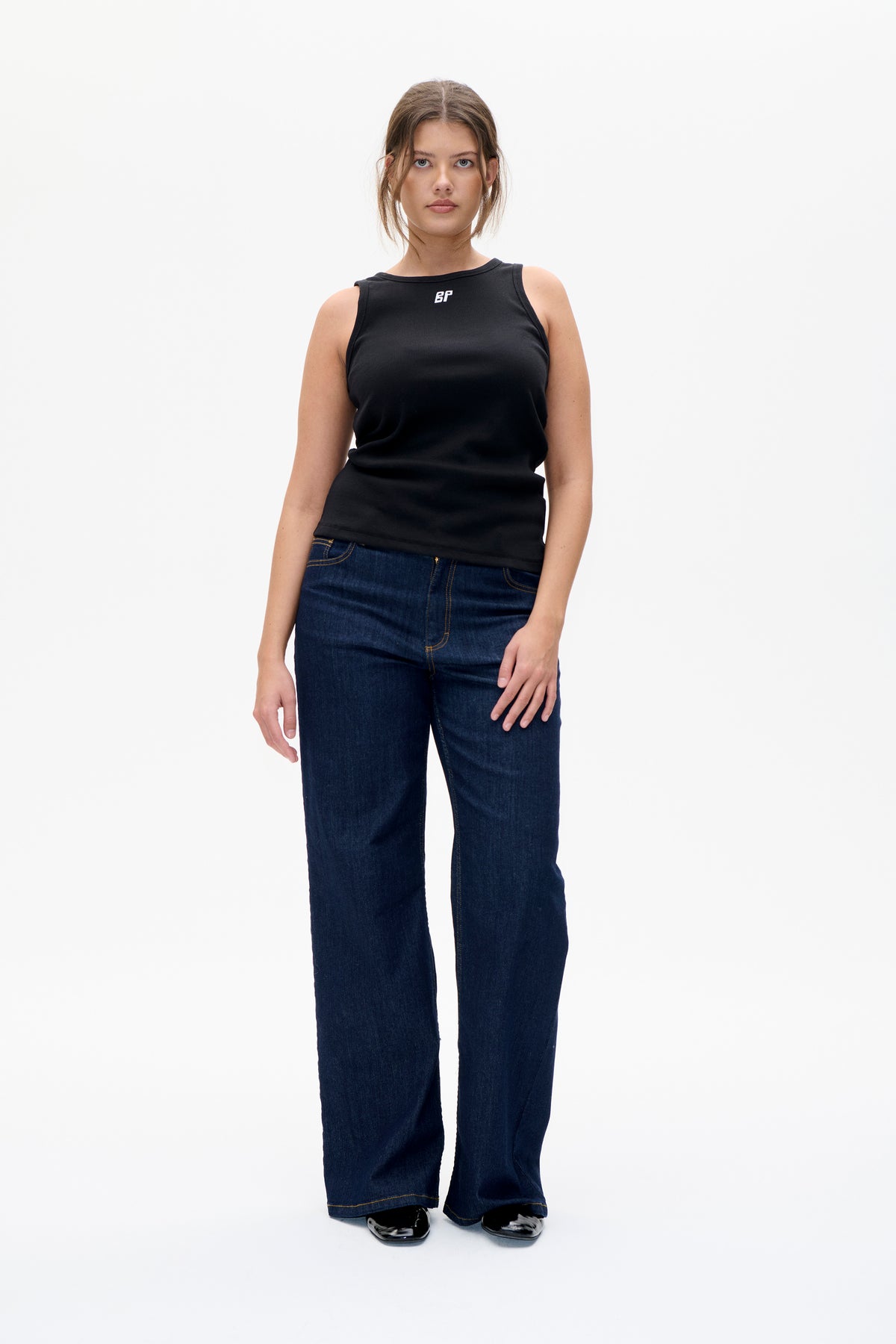 Front view of a model wearing a black sleeveless top with a small white chest logo and dark blue wide-leg jeans.