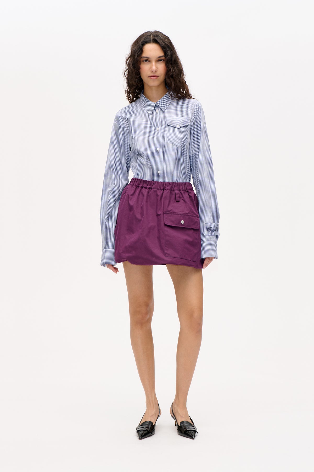 Front view of a model wearing a blue-and-white gingham button-up shirt with a chest pocket and a plum mini skirt with a large flap pocket.
