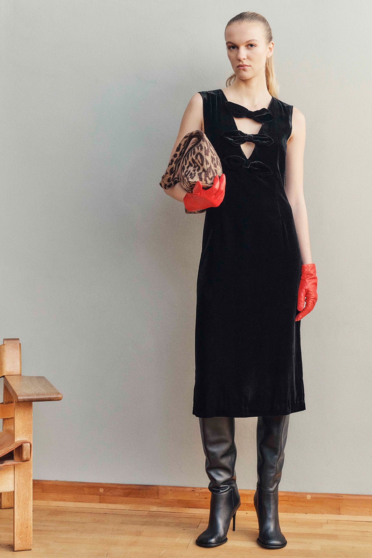 Model wearing a black sleeveless dress with bow details and front cutouts, red gloves, and tall black boots, holding a leopard-print clutch.