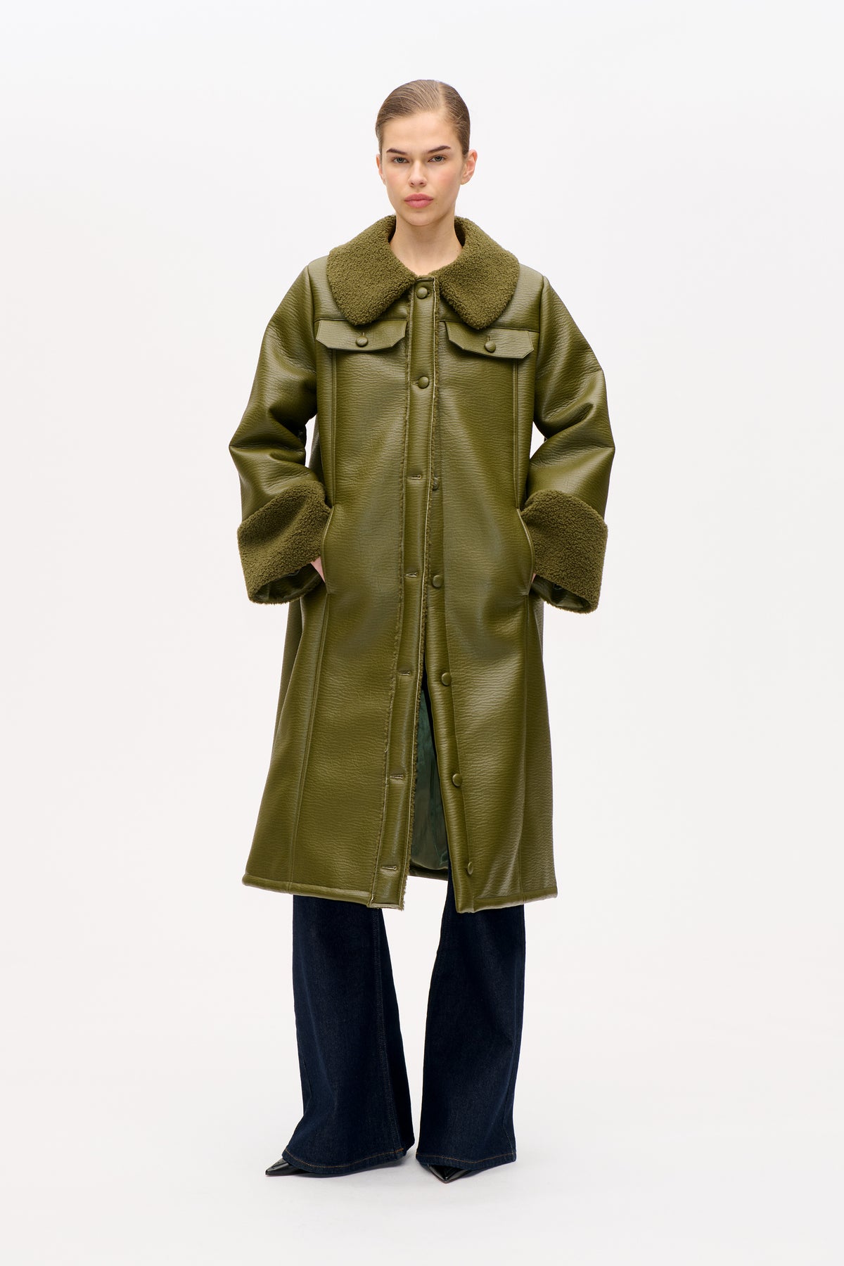 Front view of a model wearing an olive green long coat with a textured shearling-style collar and cuffs, button closure and flap chest pockets.