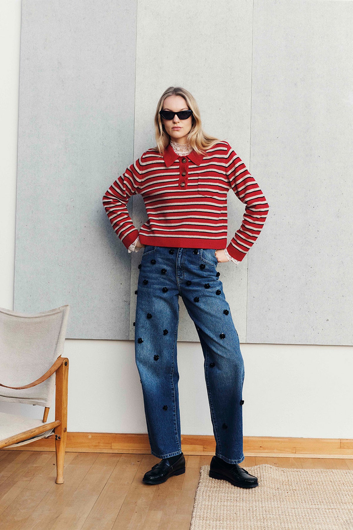 Campagneportret van een model in een gestreepte trui met knoopsluiting in rood, crème en zwart en een blauwe jeans met zwarte bloemapplicaties.