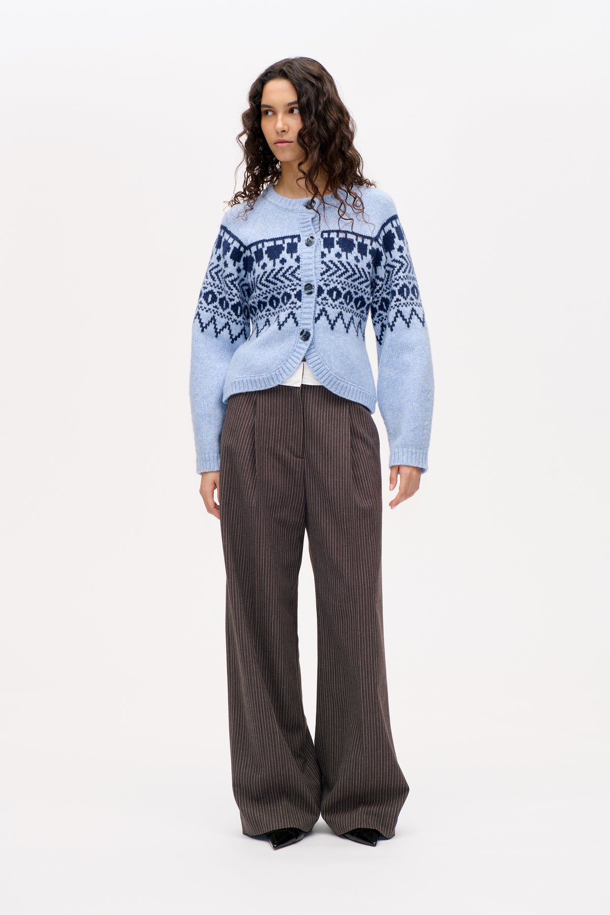 Model wearing a light blue button-front cardigan with dark navy geometric bands and brown wide-leg pinstriped trousers.