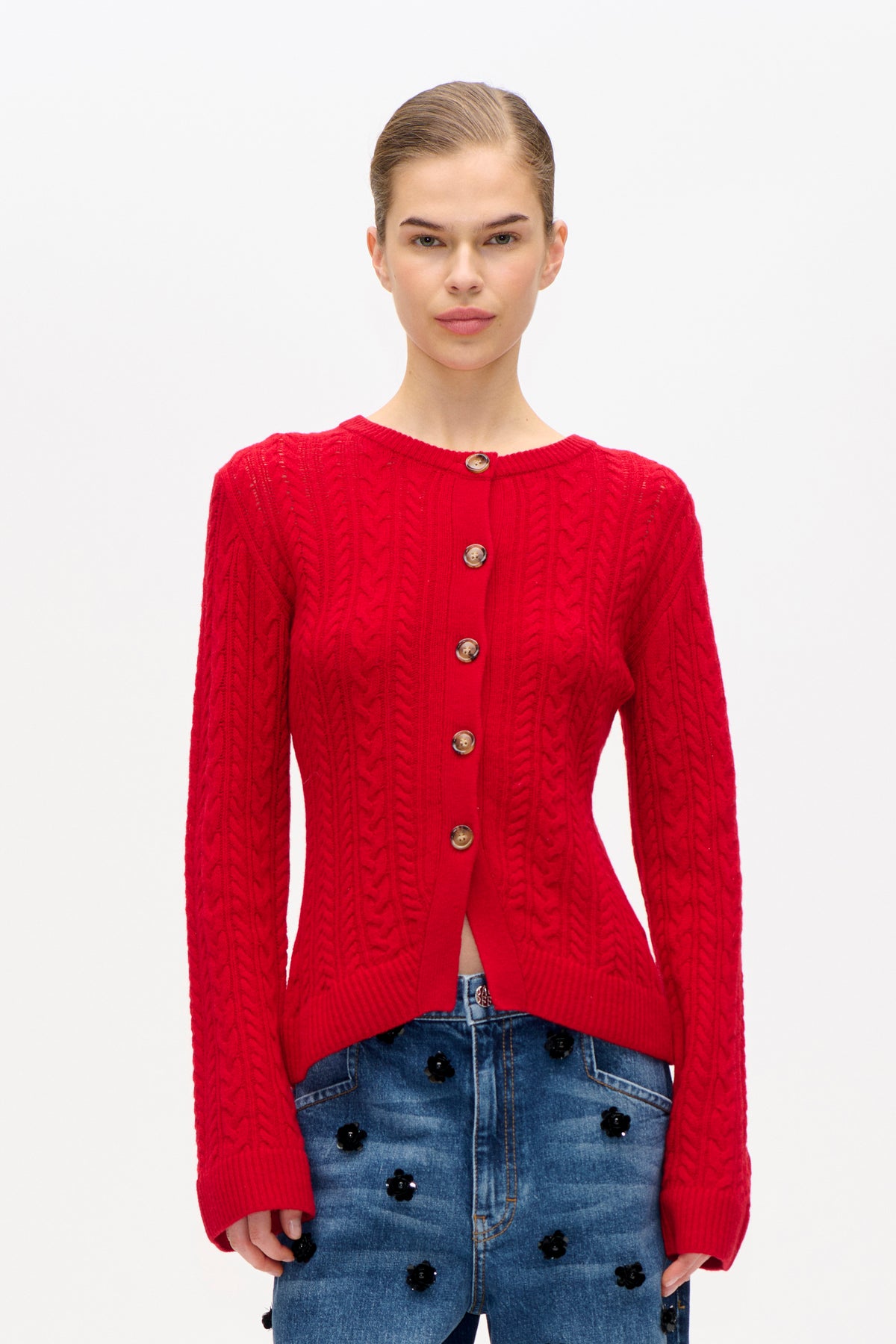 Vooraanzicht van een model dat een felrode kabelgebreide cardigan met bruine knopen draagt en een blauwe denimrok met zwarte bloemen.