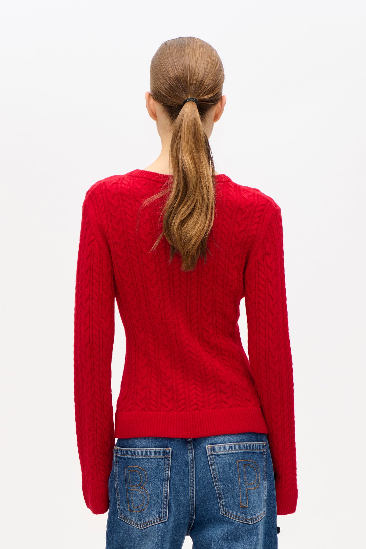 Achteraanzicht van een model in een felrode kabelgebreide trui en blauwe jeans met omlijnde letters op de achterzakken.