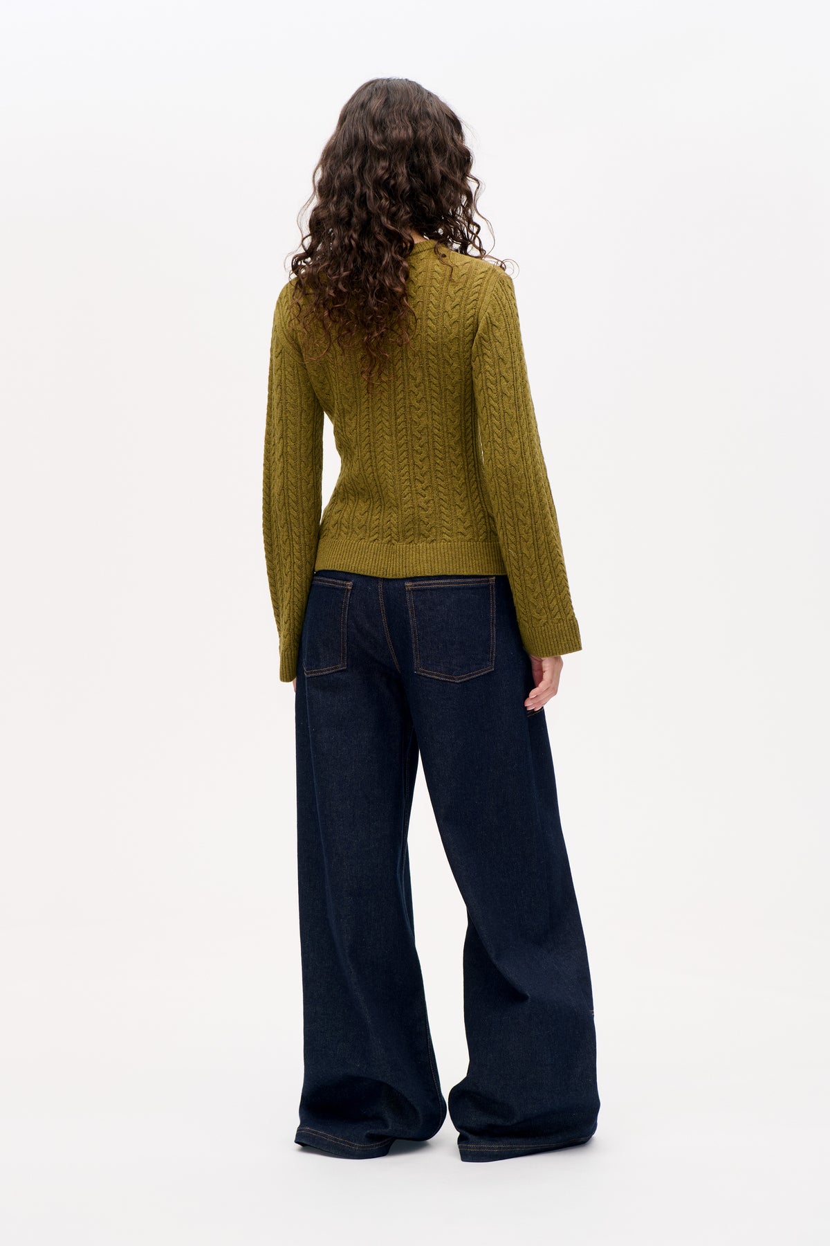 Back view of an olive green cable-knit sweater with long sleeves and ribbed hem, paired with dark wide-leg jeans.