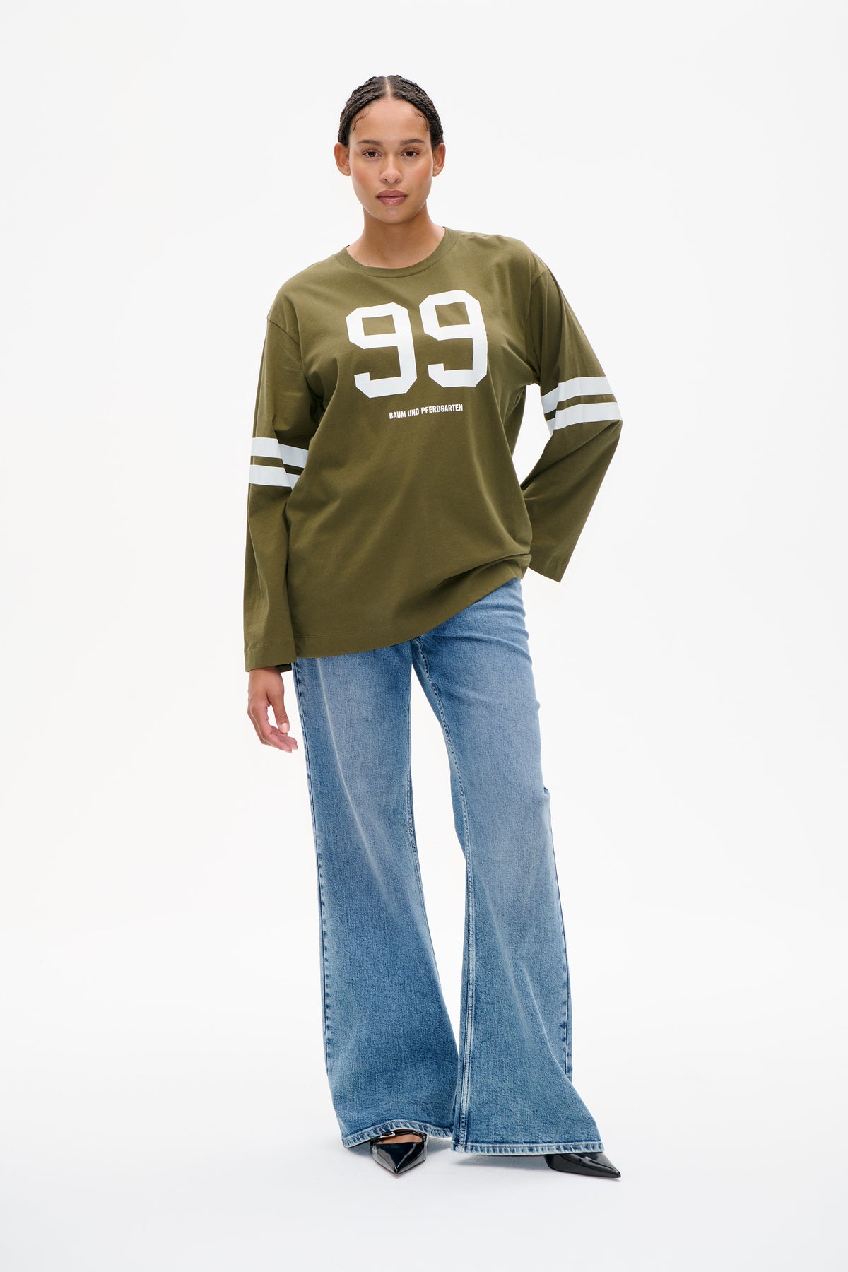 Model wearing an olive-green long-sleeve T-shirt with a large white 99 graphic and striped sleeves, paired with light-wash wide-leg jeans.