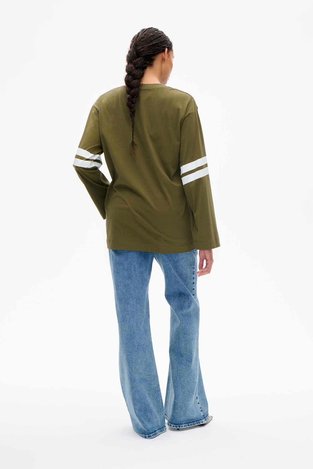 Back view of a model wearing an olive green long-sleeved top with two white stripes on each sleeve and blue jeans.