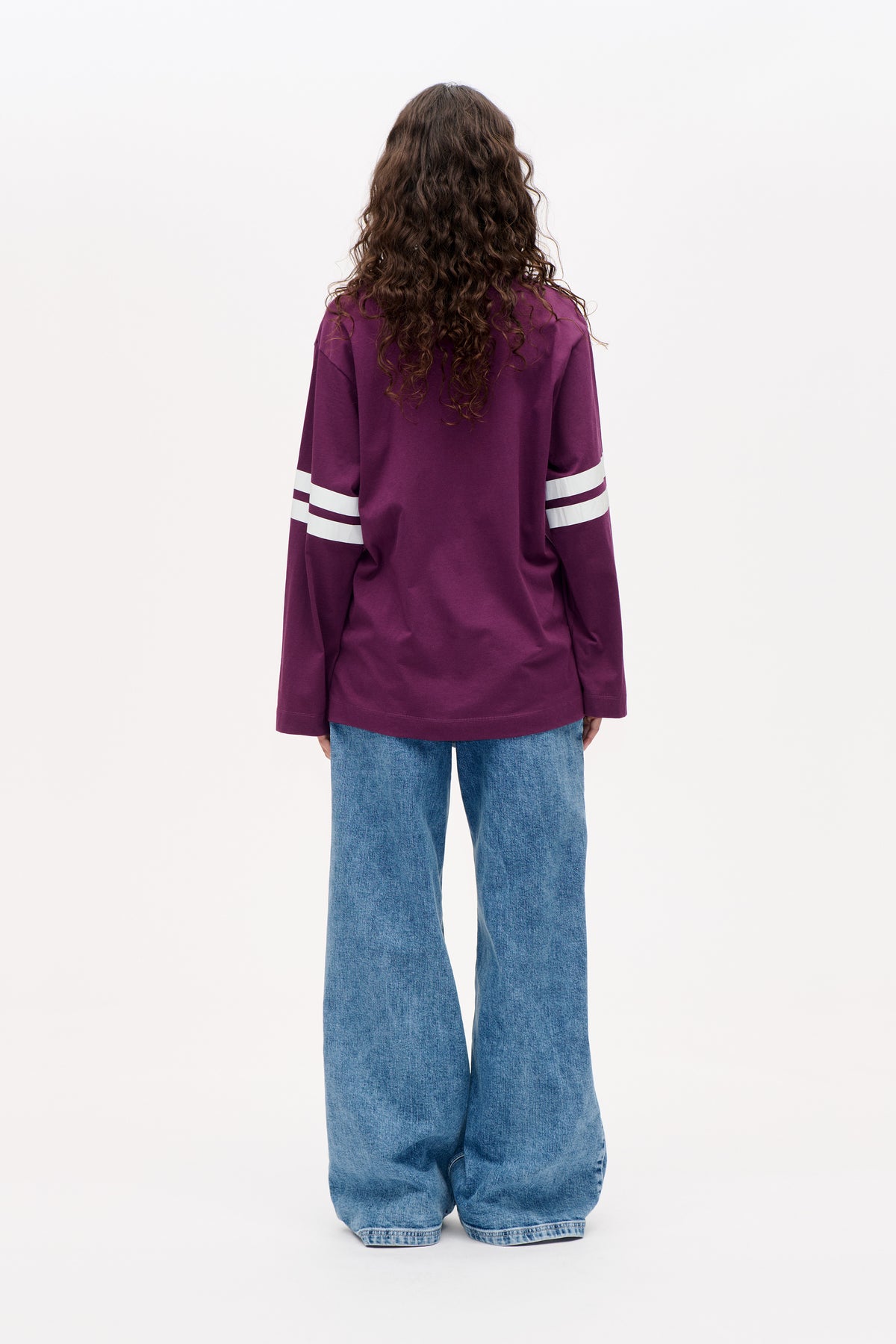 Back view of an oversized plum long-sleeve top with broad white sleeve stripes, worn with wide-leg blue jeans.