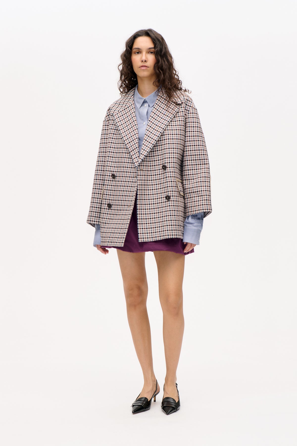 Model wearing a beige and navy checked double-breasted blazer over a blue checked shirt and purple skirt, front view.