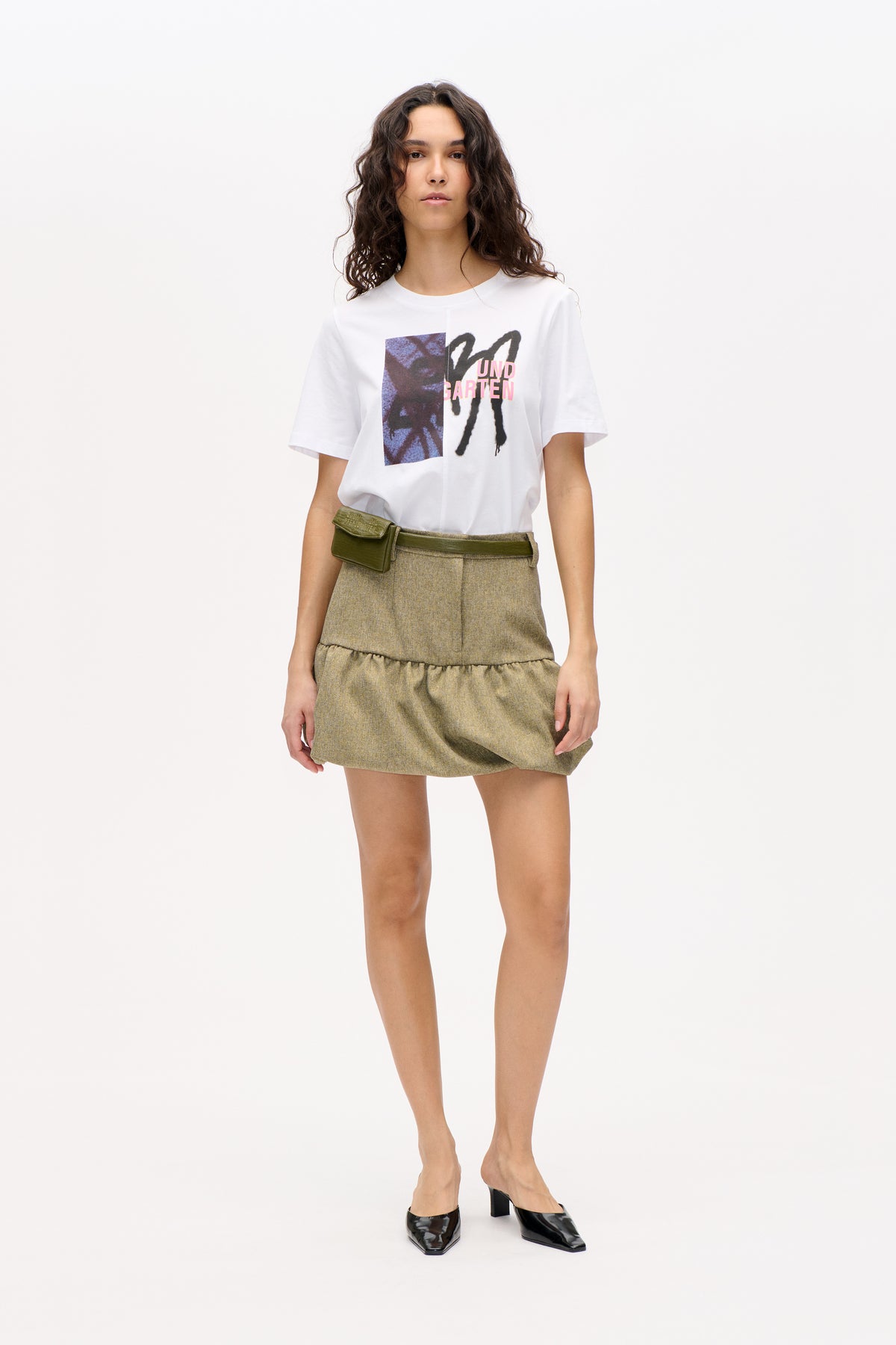 Model wearing a white short-sleeved T-shirt with a bold black, blue and pink graphic, a khaki tweed mini skirt and an olive waist bag.