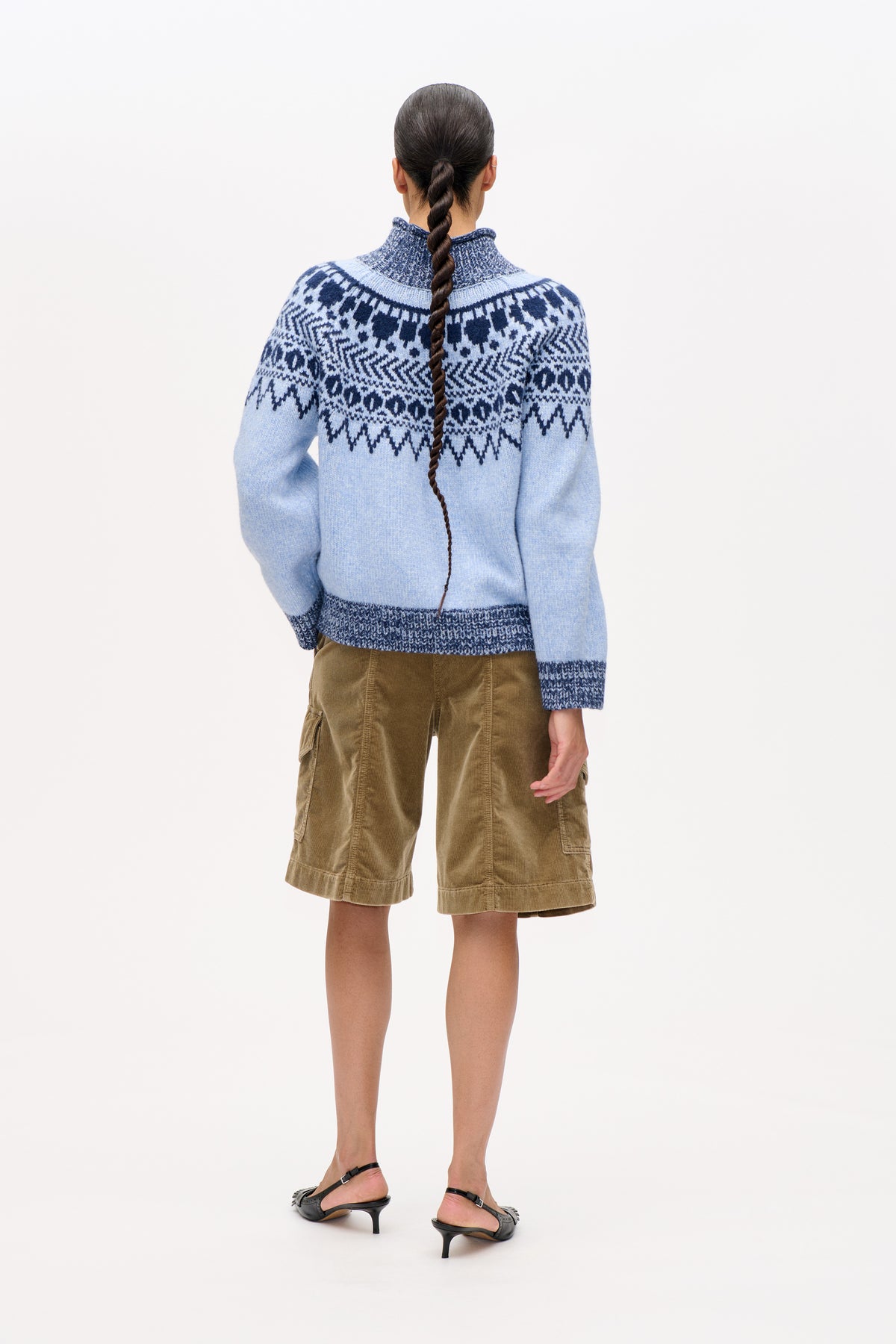 Rugaanzicht van een model dat een lichtblauwe gebreide trui draagt met een marineblauw geometrisch motief op het schouderstuk en een bruine ribfluwelen korte broek.
