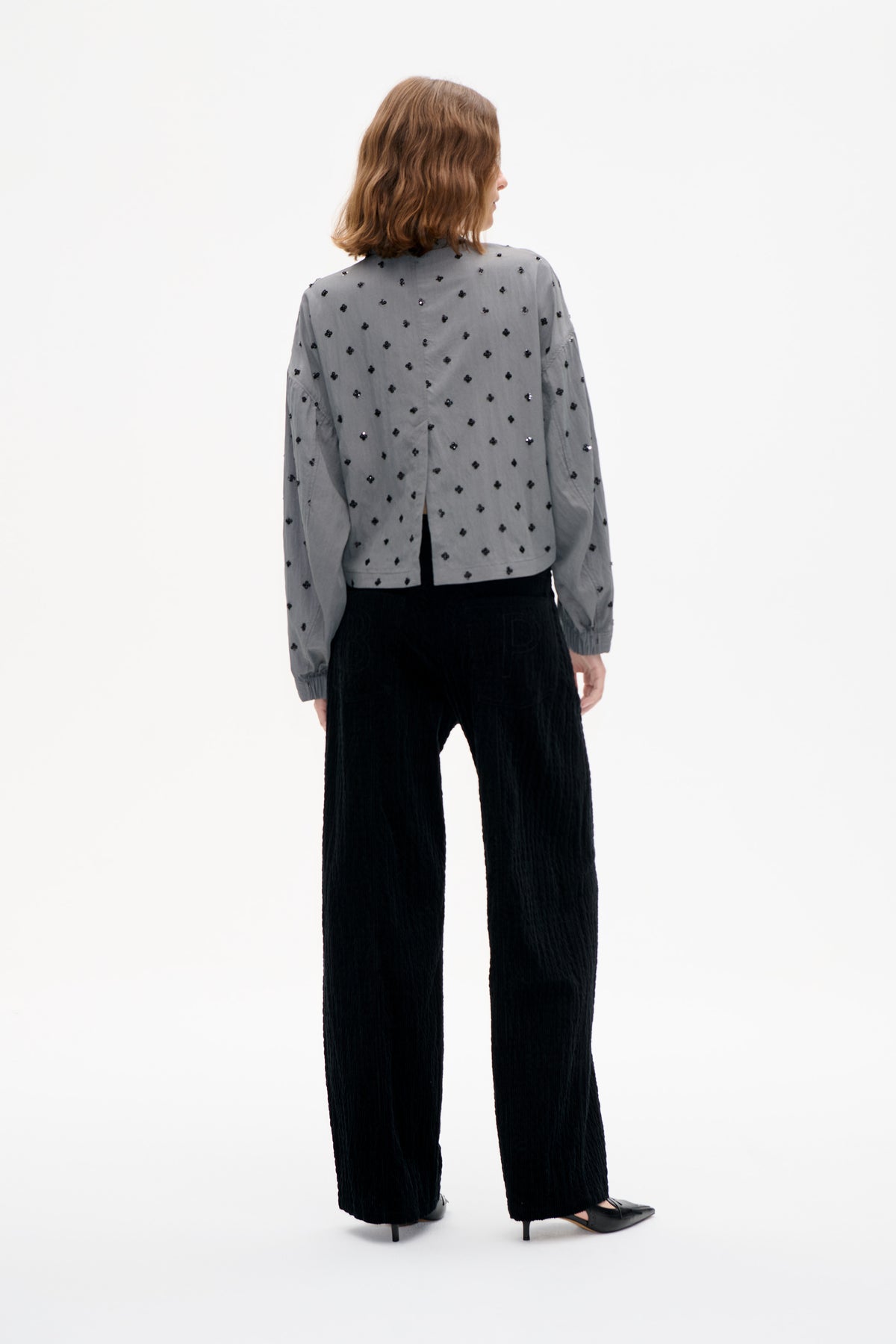 Back view of a model wearing a grey long-sleeved blouse with scattered black embellishments and black wide-leg trousers.