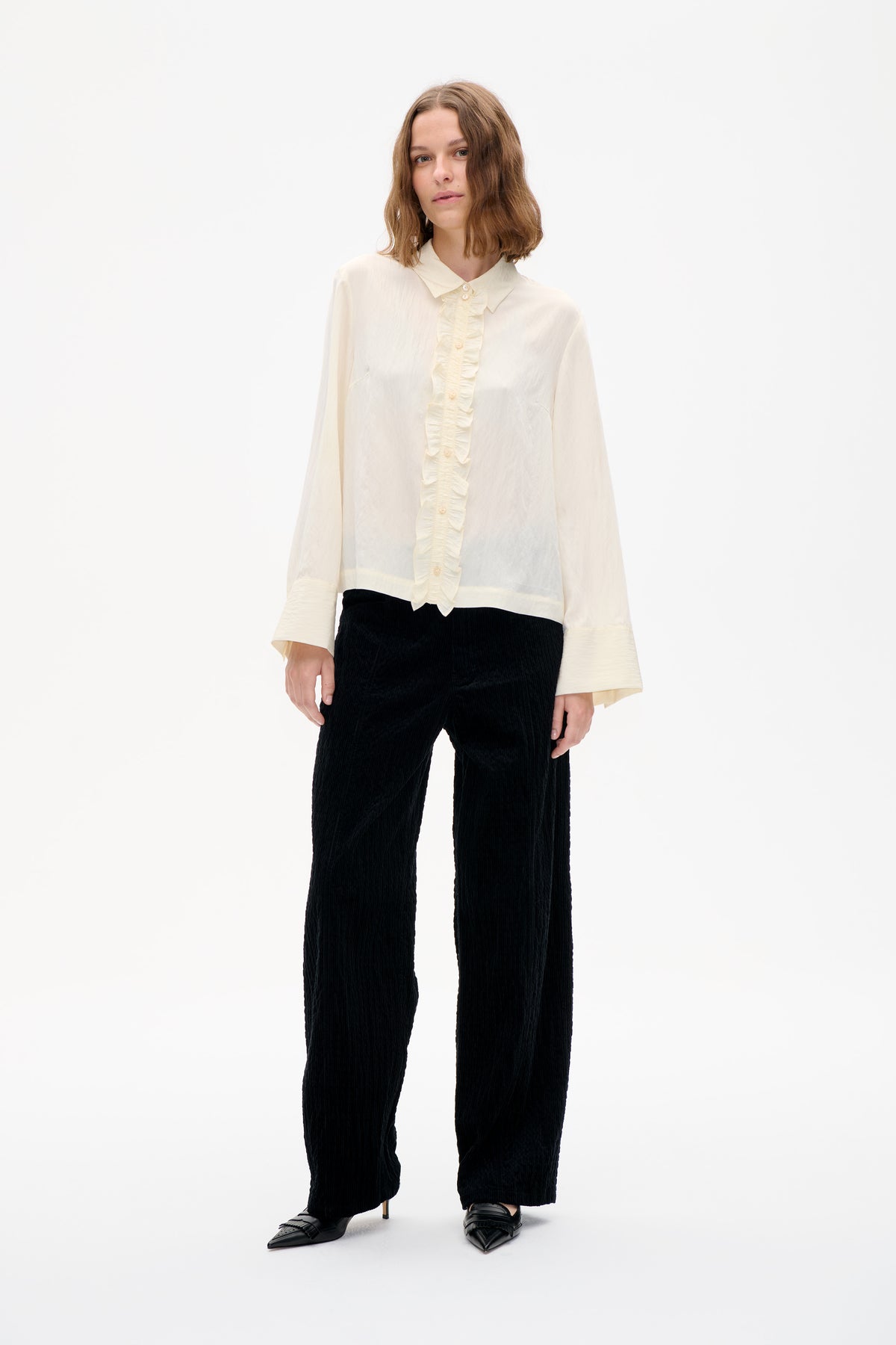 Model trägt eine elfenbeinfarbene, langärmelige Bluse mit spitzem Kragen und rüschiger Knopfleiste sowie eine schwarze, strukturierte, weit geschnittene Hose.