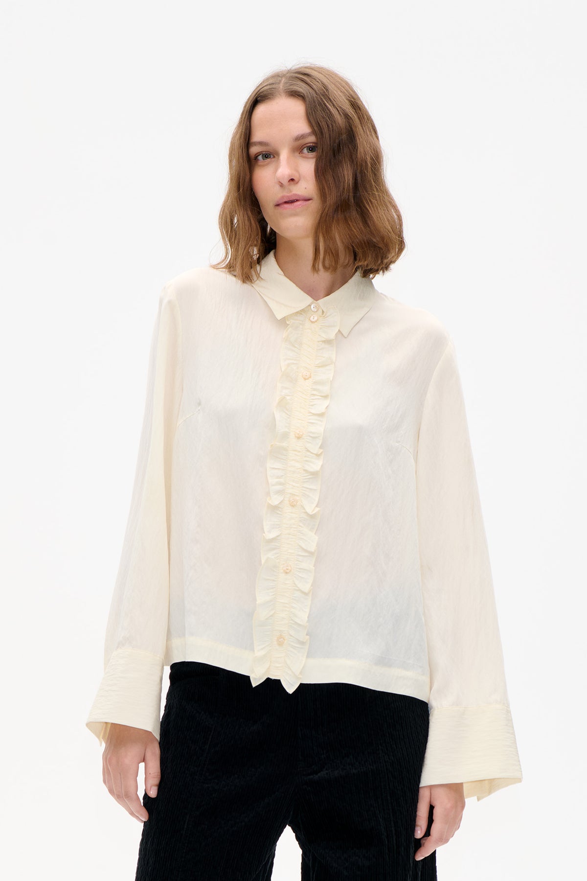 Front detail of an ivory blouse with a pointed collar, buttons and a gathered ruffle placket, styled with black trousers.