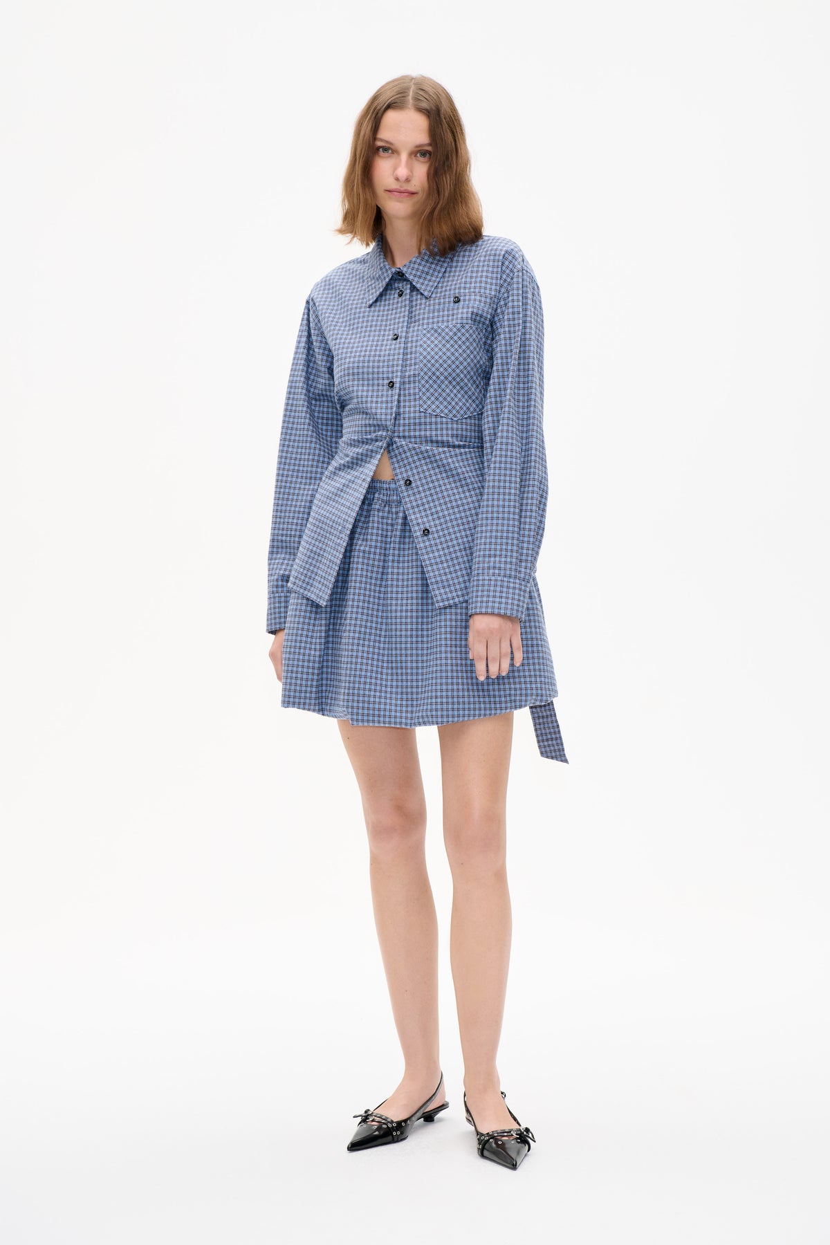 Model draagt een blauw geruite blouse met knoopsluiting en borstzak, gecombineerd met een bijpassende geplooide rok.