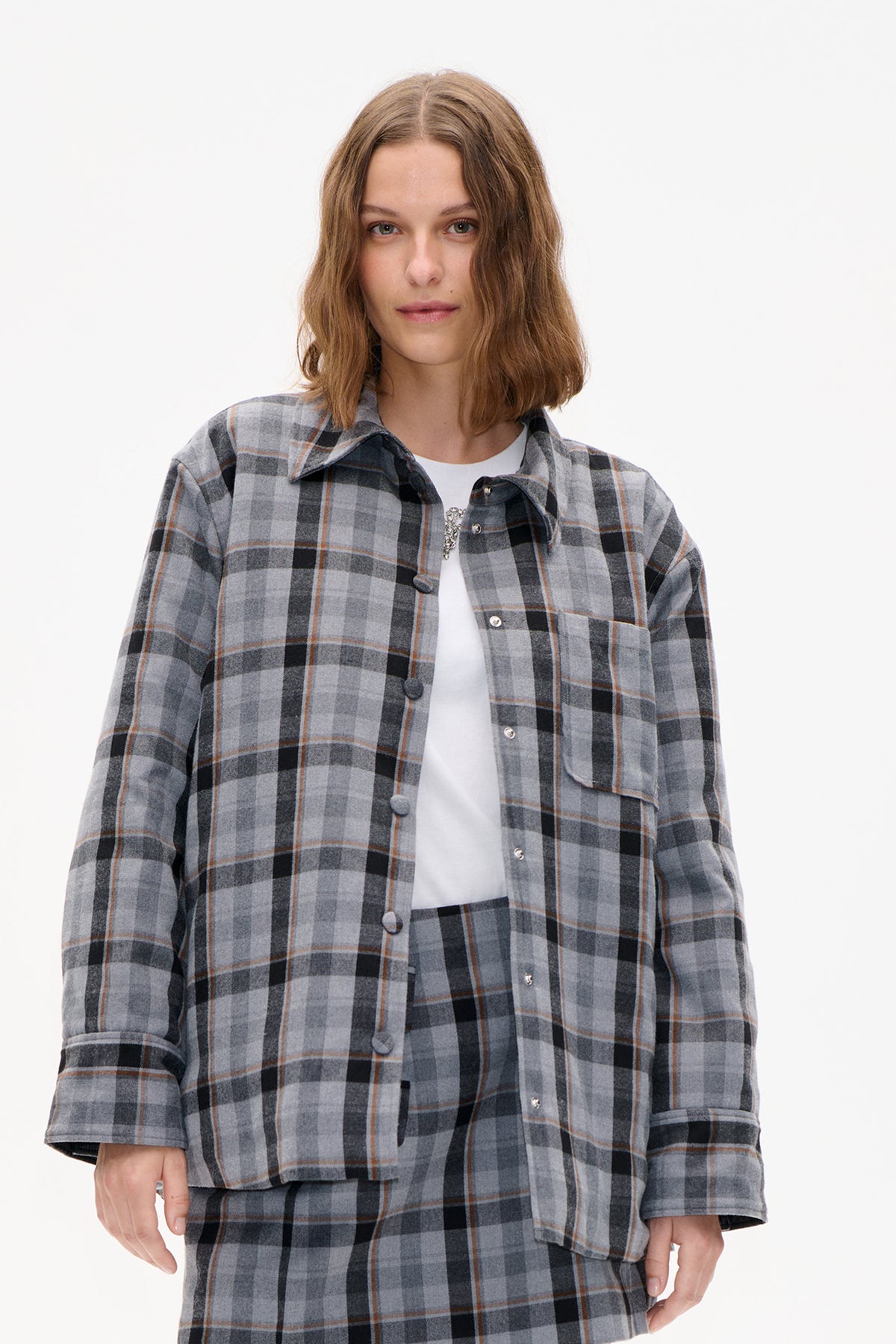 Front view of a model wearing a grey plaid button-front shirt jacket over a white top, with a chest pocket and silver buttons.