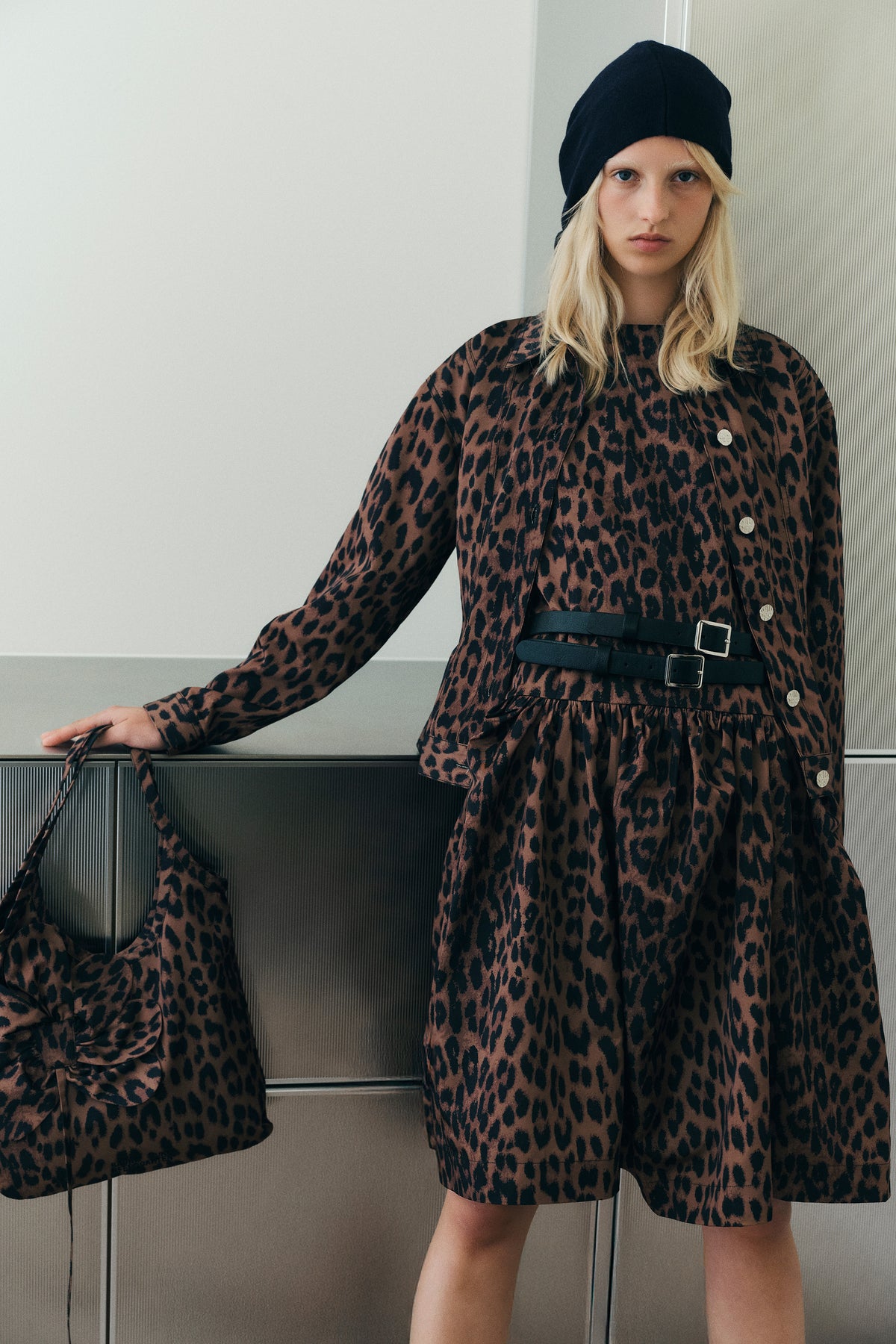 Model trägt ein braun-schwarzes, leopardbedrucktes Hemdkleid mit Gürtel und eine schwarze Beanie. Sie hält ein passendes Kleidungsstück vor einem hellen Innenraumhintergrund.