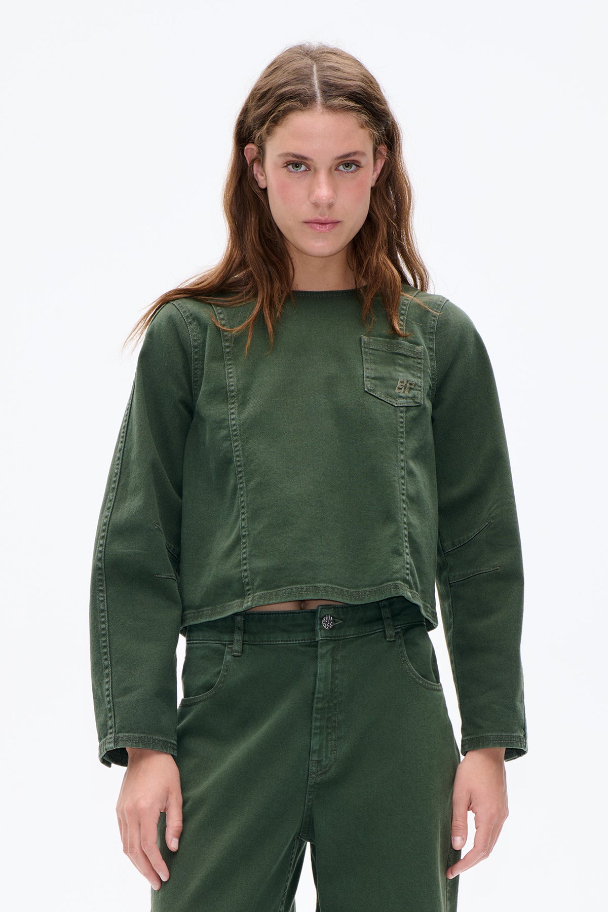 Front view of a model wearing a cropped olive-green long-sleeved top with contrast seams and a chest patch pocket, paired with matching trousers.