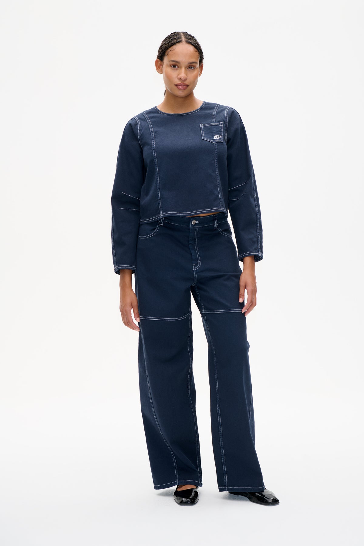 Model wearing a navy long-sleeved top with contrast topstitching and chest pocket, paired with wide-leg trousers.