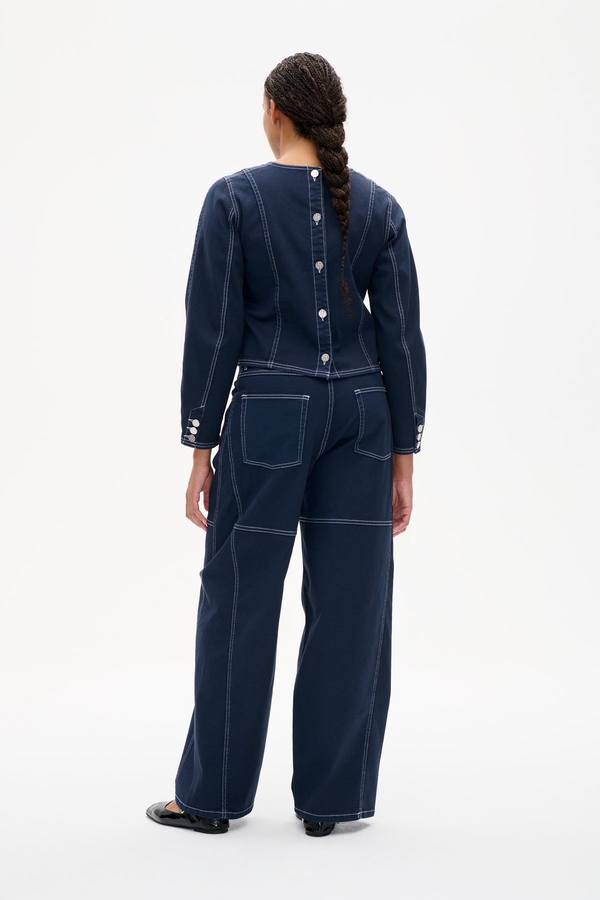 Back view of a model wearing a dark indigo denim jacket with contrast stitching and silver buttons, paired with matching wide-leg trousers.