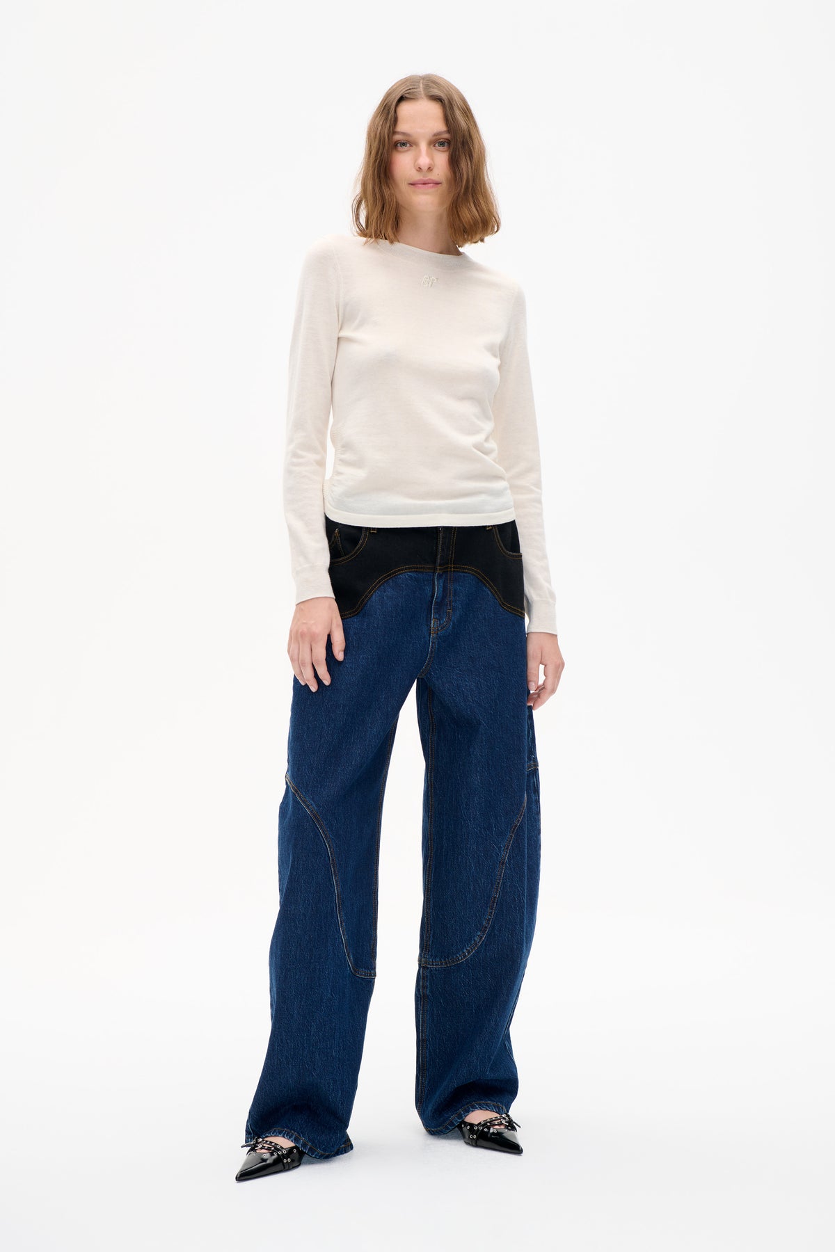 Front view of a model wearing an ivory long-sleeved crew-neck top with a small chest monogram and wide-leg dark blue jeans.