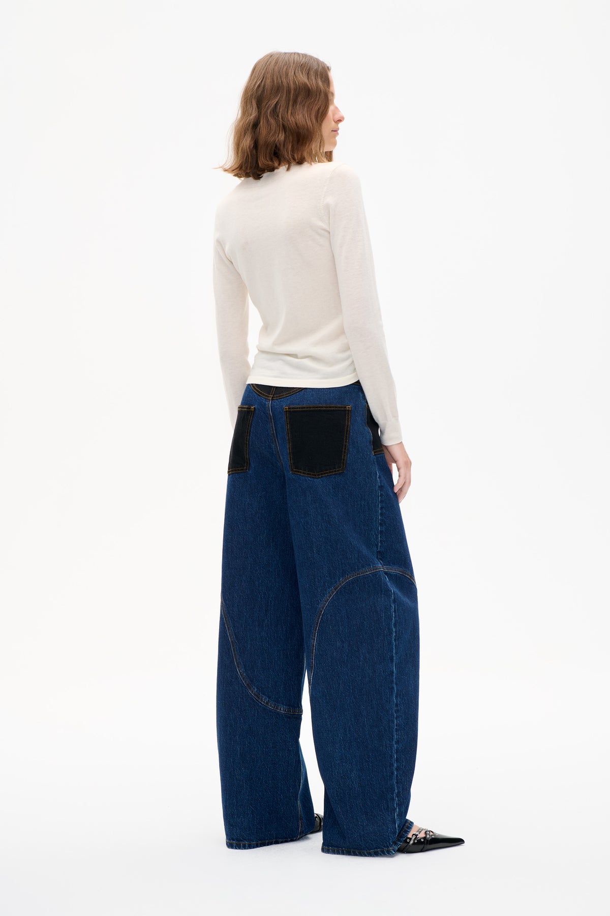 Model shown from the back wearing an off-white long-sleeved top and wide-leg dark blue jeans with contrasting patch pockets and curved seams.