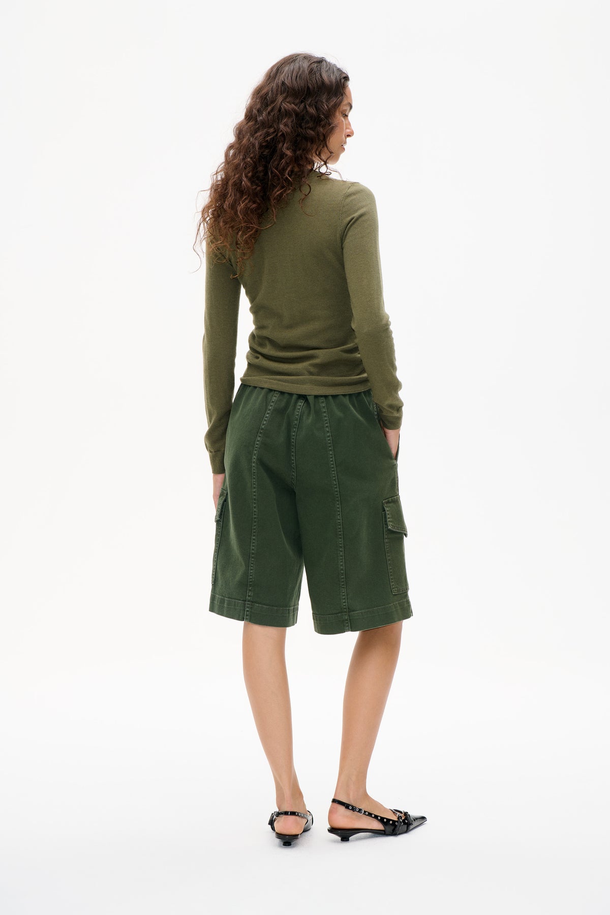 Model turned in profile wearing an olive long-sleeve top, dark green knee-length cargo shorts and black studded slingback flats.