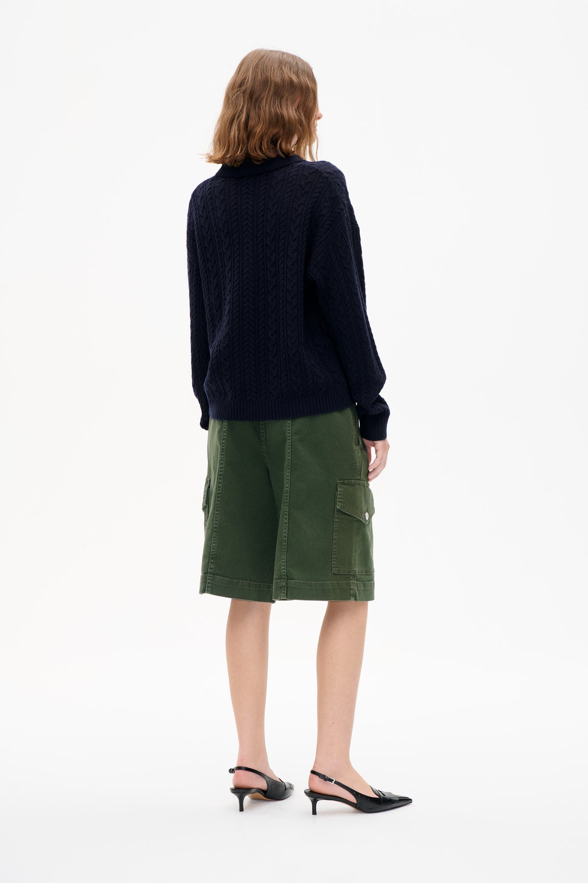 Rear view of a model wearing a dark navy cable-knit sweater with a ribbed collar and an olive green cargo skirt.