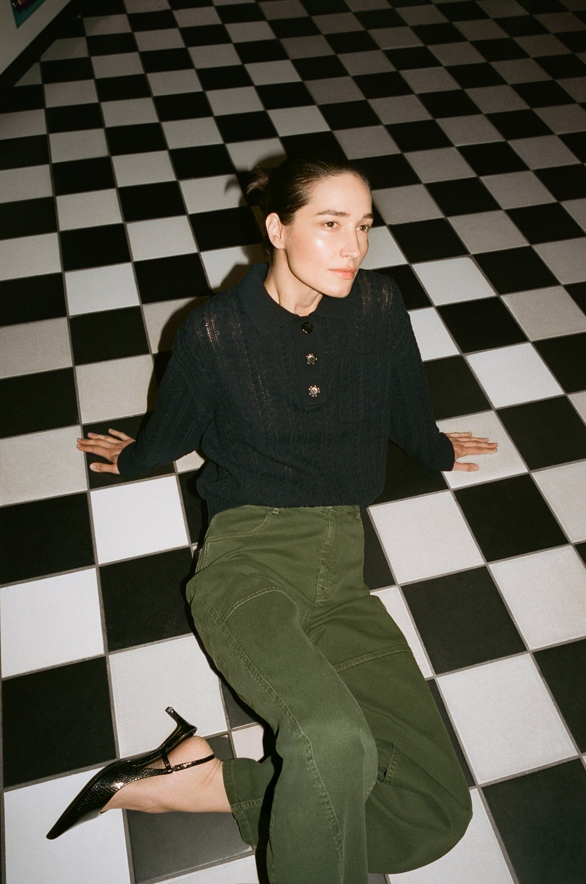 A seated model wears a dark navy textured button-front sweater and olive green trousers against a black-and-white checkerboard floor.