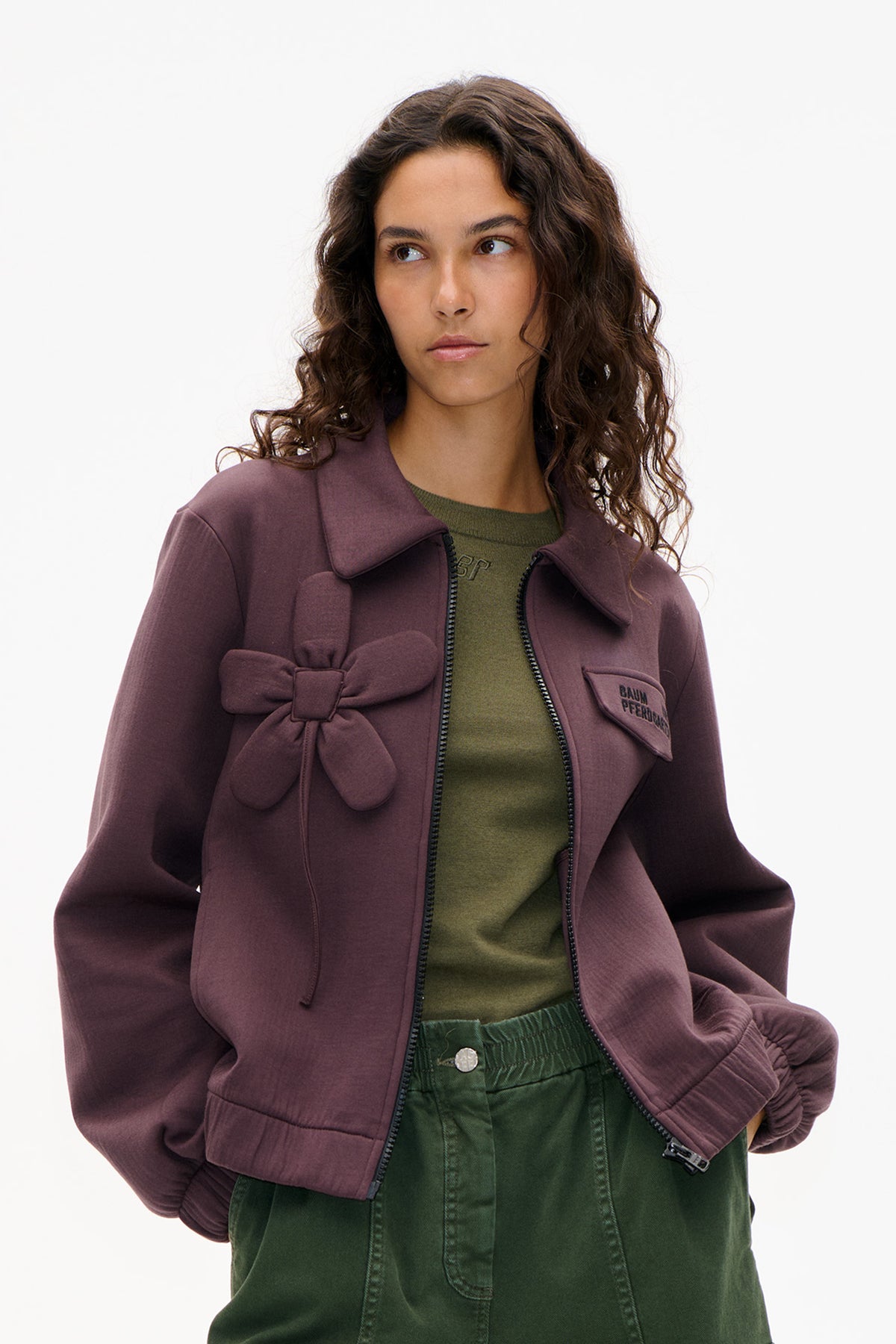 Model wearing a mauve zip-front jacket with oversized collar, chest bow and gathered cuffs over a green top and trousers.